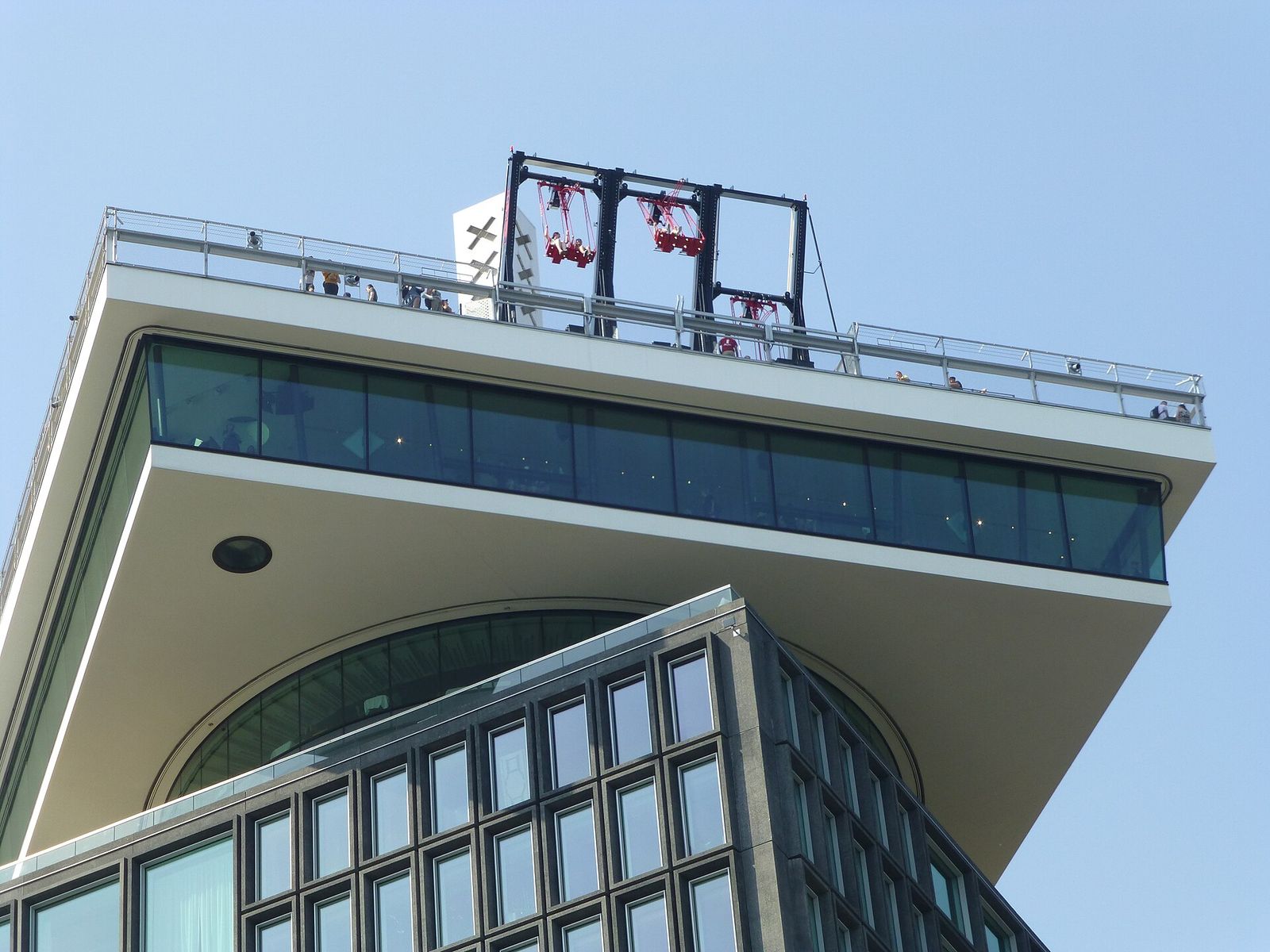 The cantilevered viewing deck on top of the A'DAM Tower seen from below, with the red seats of the swing hanging over the edge against a blue sky