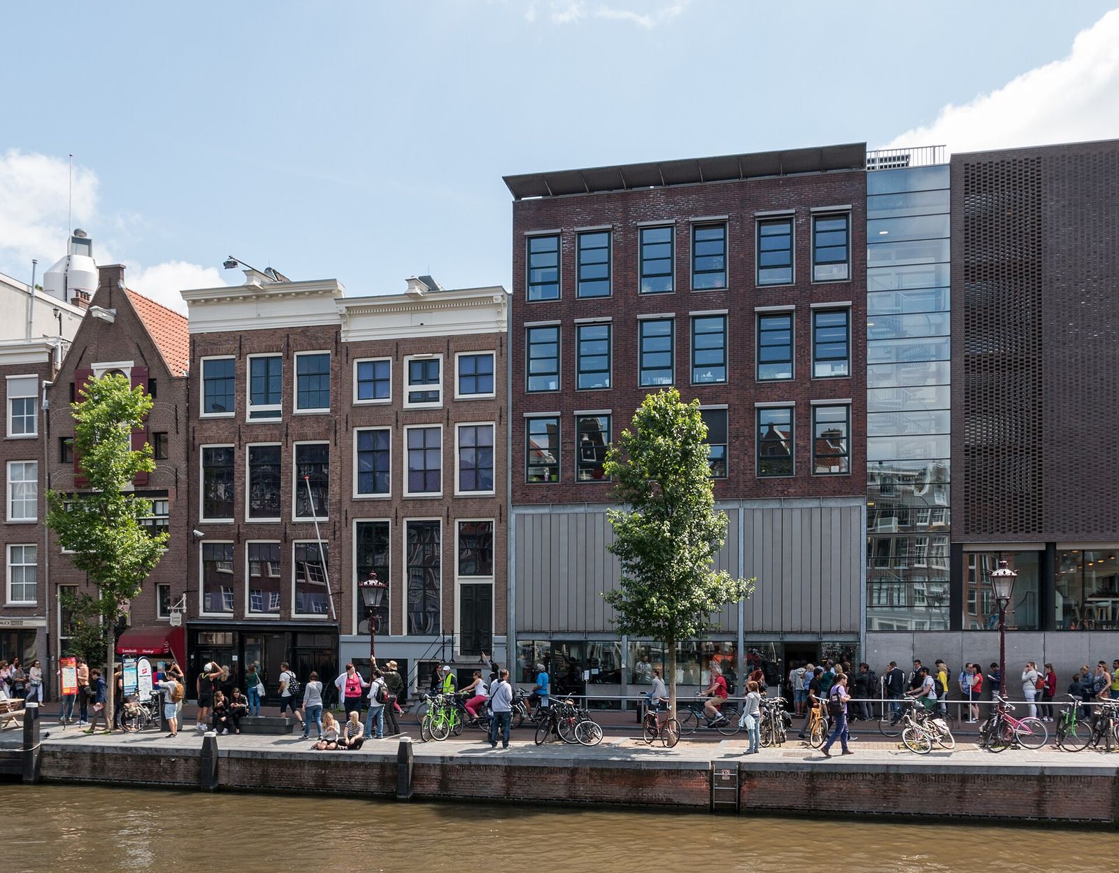 The Anne Frank House on the Prinsengracht, a narrow canal house next to the museum's modern brick-and-glass building, with visitors and parked bikes along the canal on a sunny day