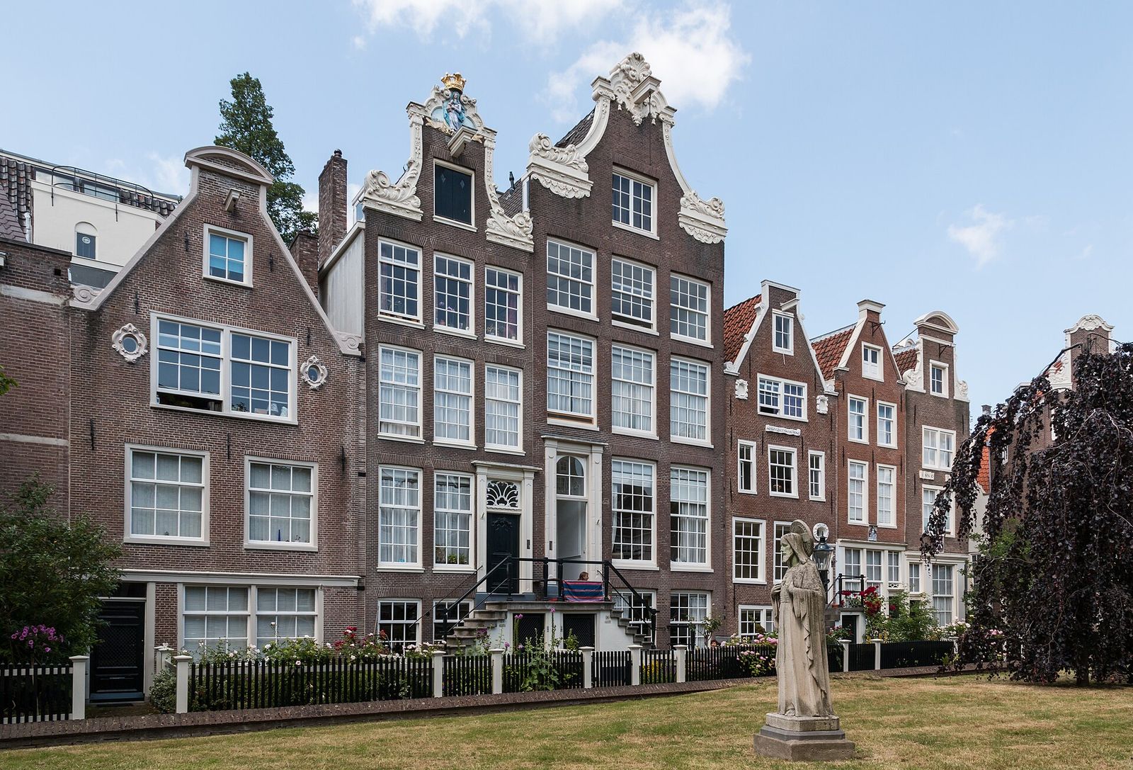 Tall brick almshouses with white window frames and ornate gables around the lawn of the Begijnhof, with a statue in the garden under a blue sky
