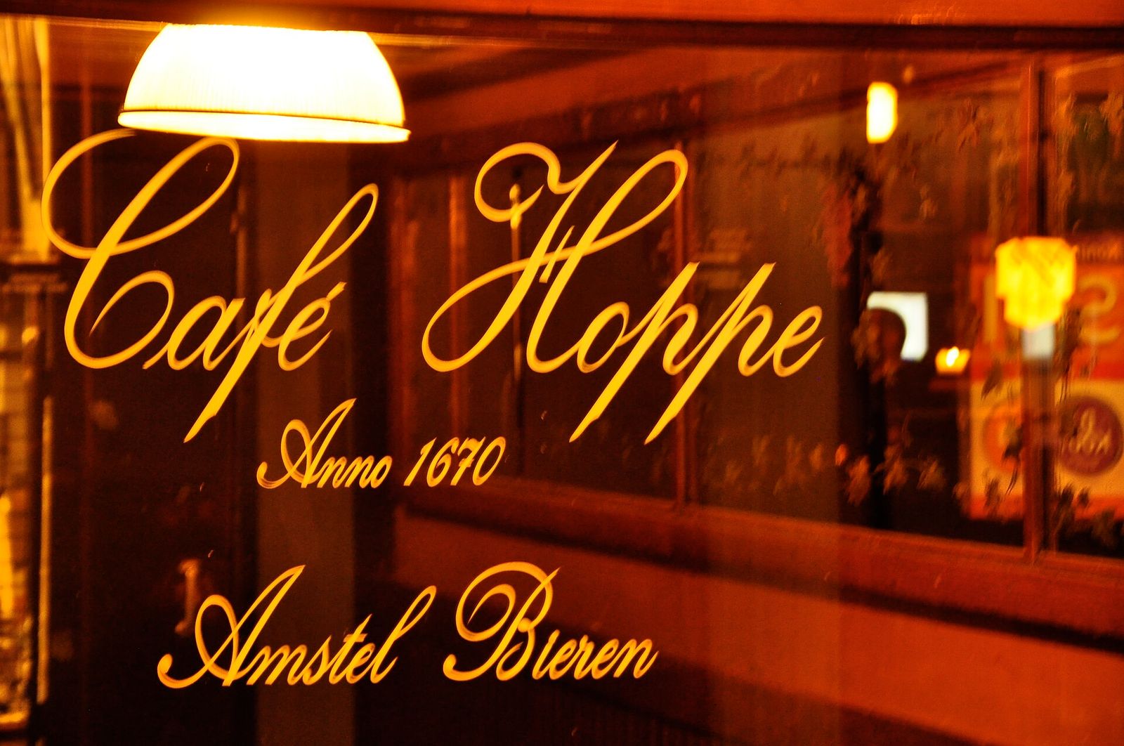 Gold lettering reading Café Hoppe, Anno 1670, Amstel Bieren on the café's window, lit warmly from inside