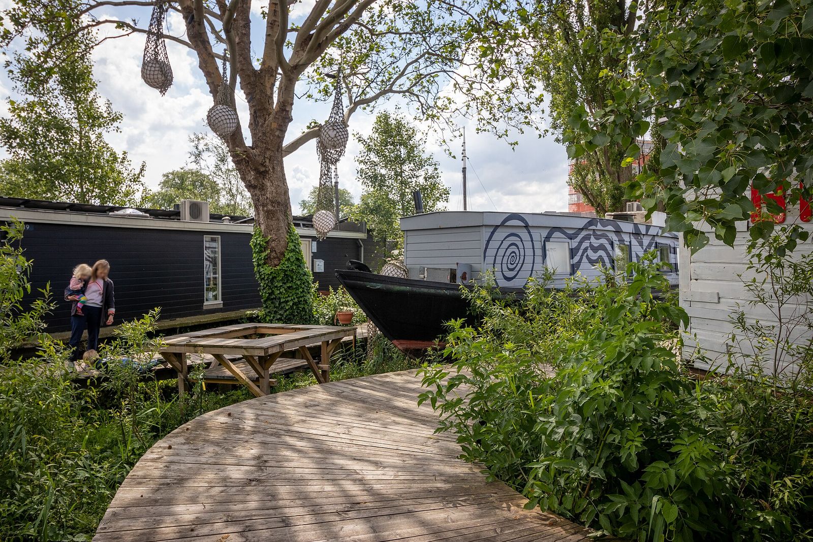 A winding wooden boardwalk through tall green plants past dark-painted houseboats resting on land at De Ceuvel