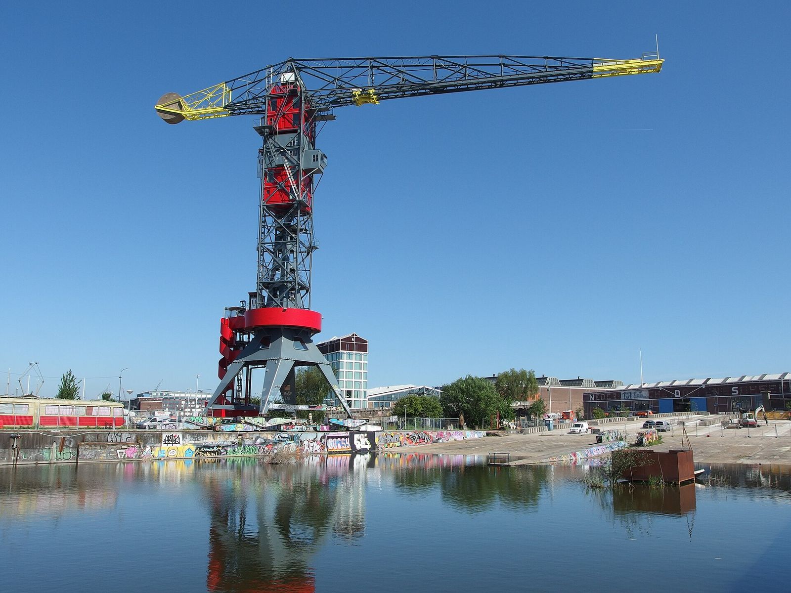 The red-and-grey Faralda harbor crane at the NDSM wharf, reflected in still water, with old shipyard buildings behind