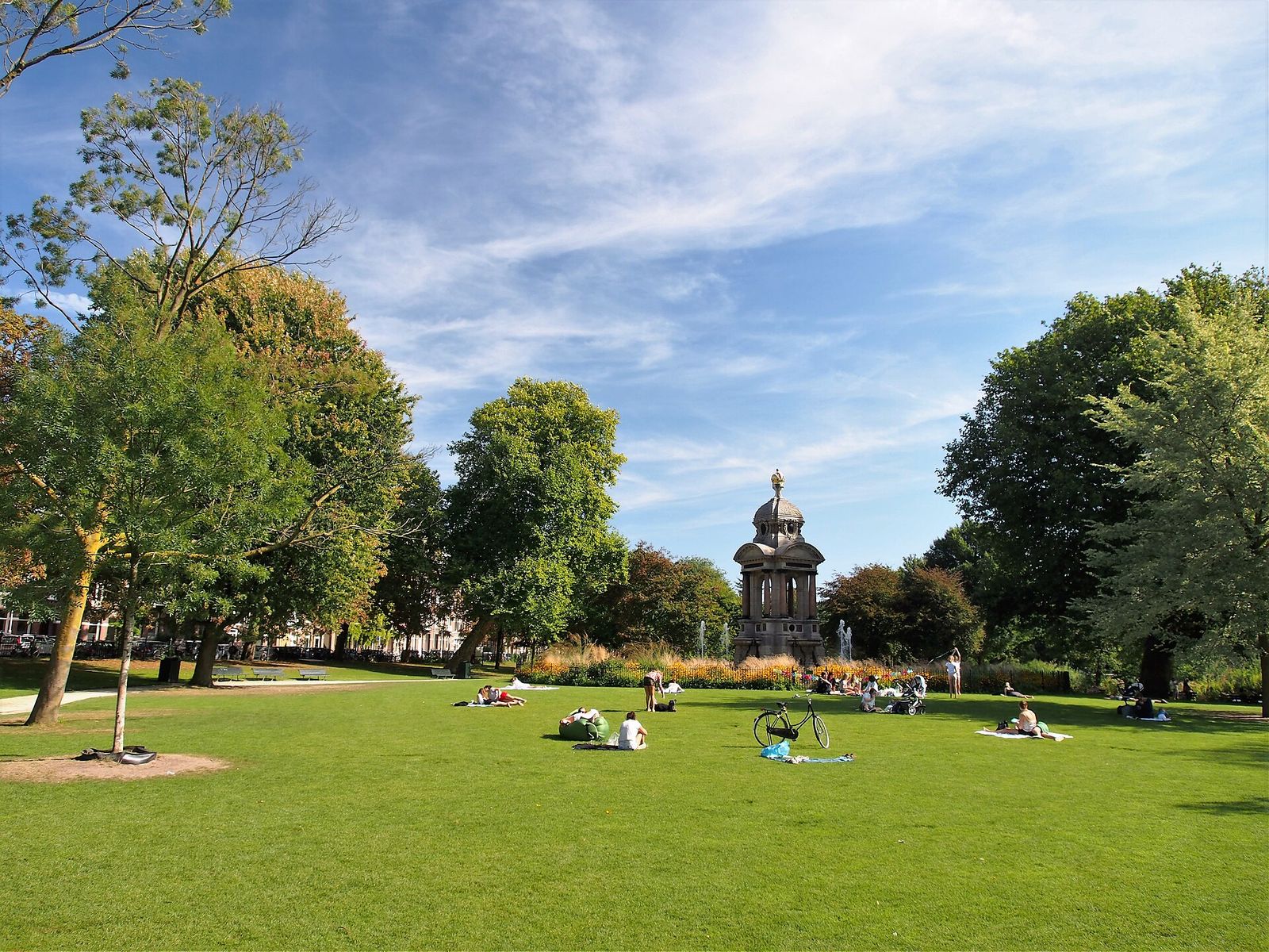 People sitting on the lawn of the Sarphatipark on a sunny day, with the Sarphati monument between the trees