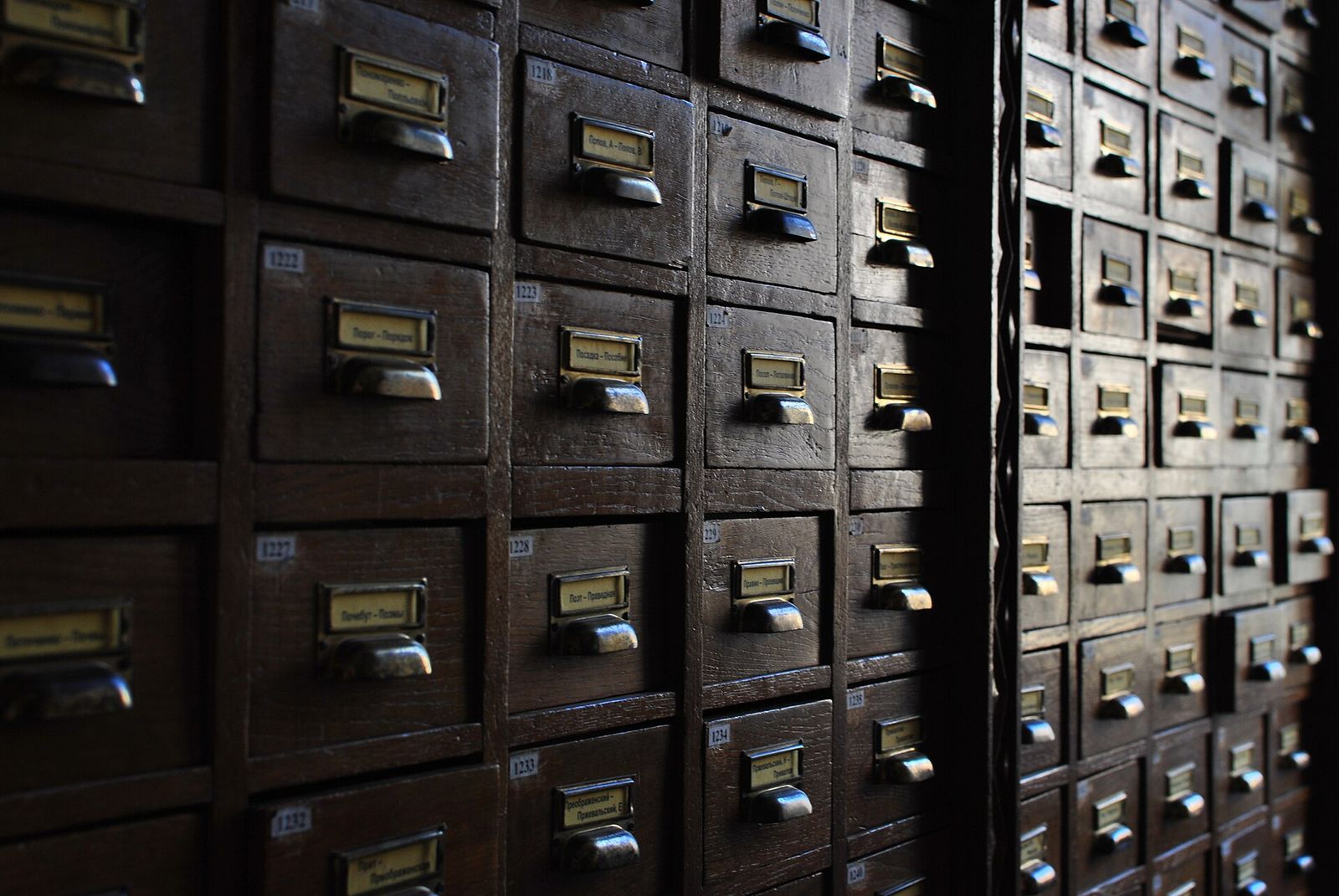 Rows of dark wooden card catalogue drawers with brass handles, each holding a small label with a range of Cyrillic headings