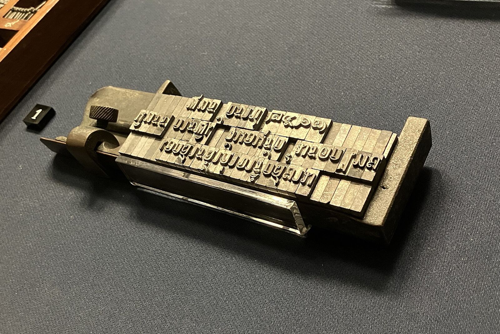 Pieces of metal type for Thai script set in lines on a brass composing stick, displayed on grey cloth in a museum case