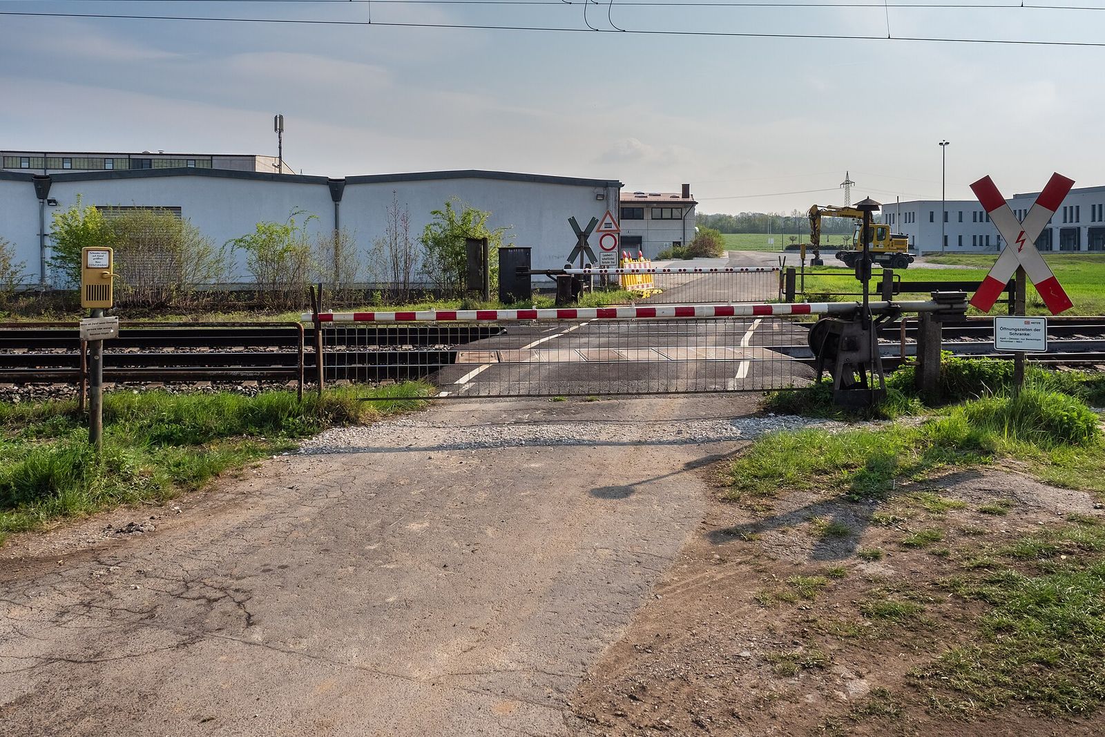 A red and white barrier lowered across a road at a railway level crossing, with a red and white St Andrew's cross sign on the right and low industrial buildings behind