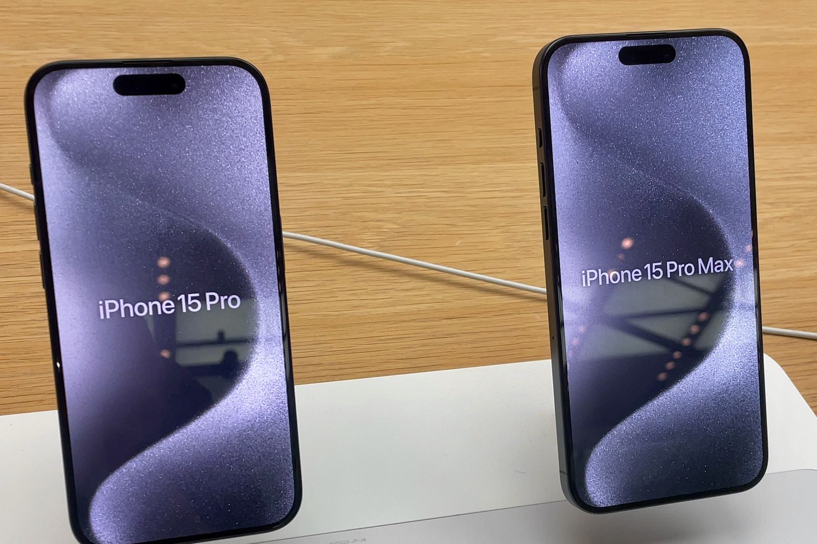 Two iPhones standing on a store display table, their screens reading iPhone 15 Pro and iPhone 15 Pro Max, the Pro Max slightly larger