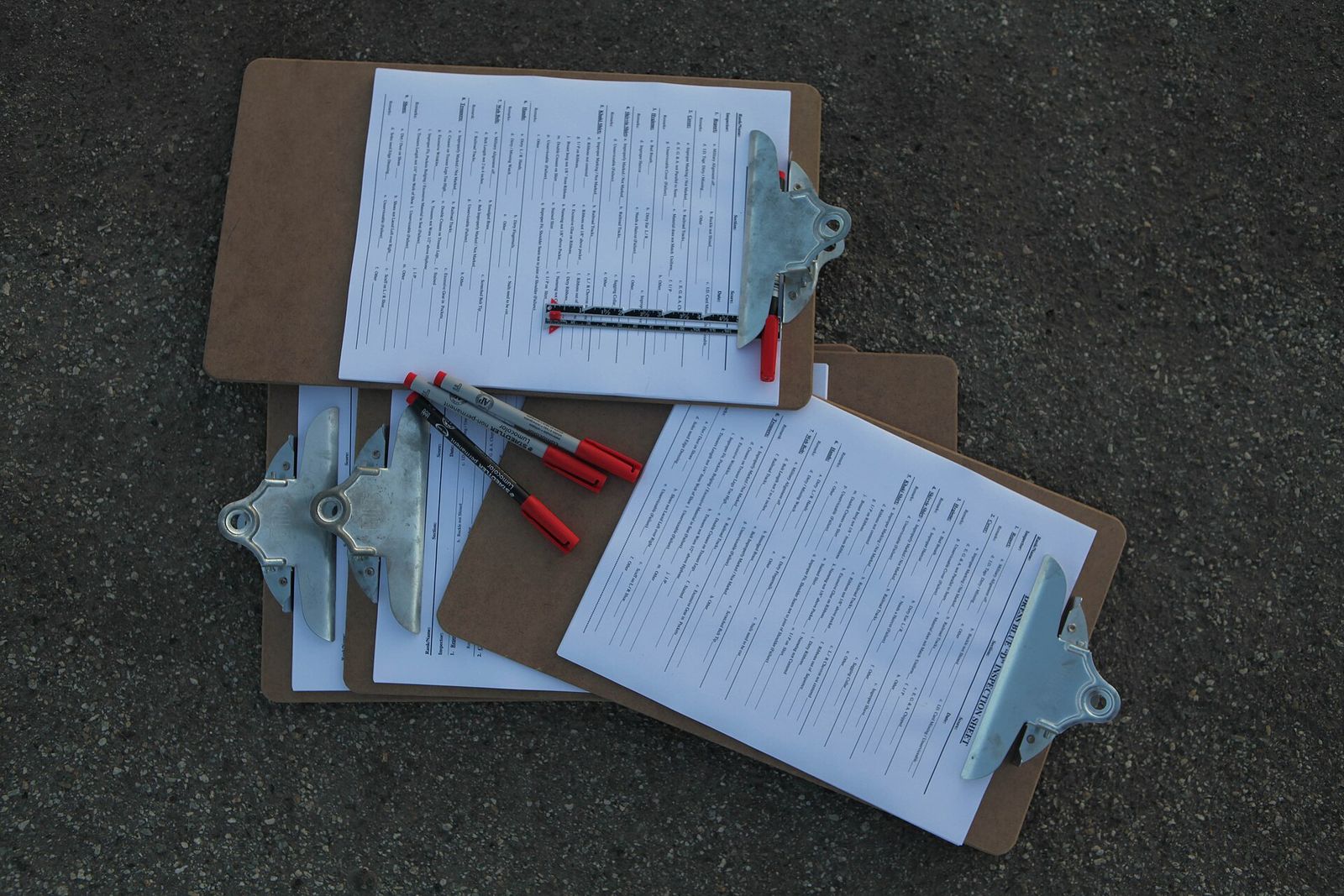Four clipboards lying on dark asphalt, each holding a printed inspection checklist, with three felt-tip pens with red caps across them