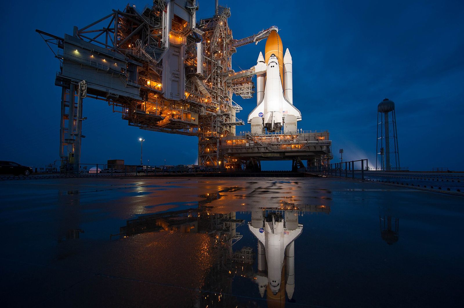 Space Shuttle Atlantis standing on its launch pad after dark, lit up beside the steel service tower, with the whole scene reflected in rainwater on the concrete