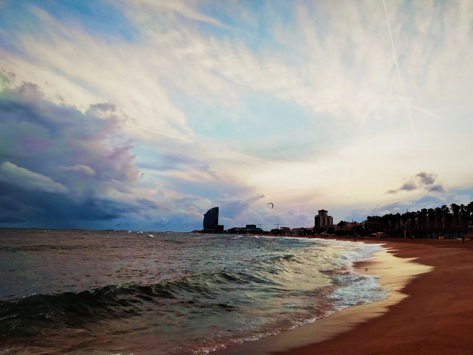 Barceloneta beach at dusk seen from a breakwater: waves running up the sand toward a line of palms, with the sail-shaped W hotel on the horizon under streaky clouds