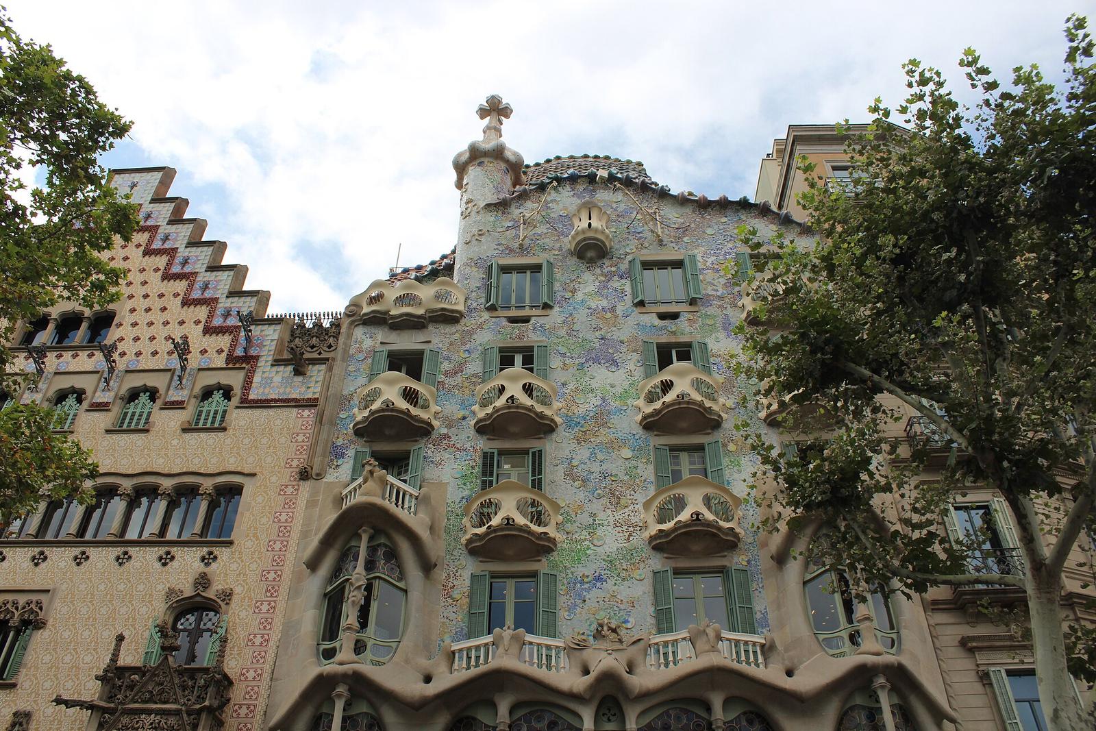 Casa Amatller's stepped, tiled gable beside the blue-green mosaic façade of Casa Batlló, with its mask-like balconies and cross-topped turret, seen from Passeig de Gràcia between plane trees