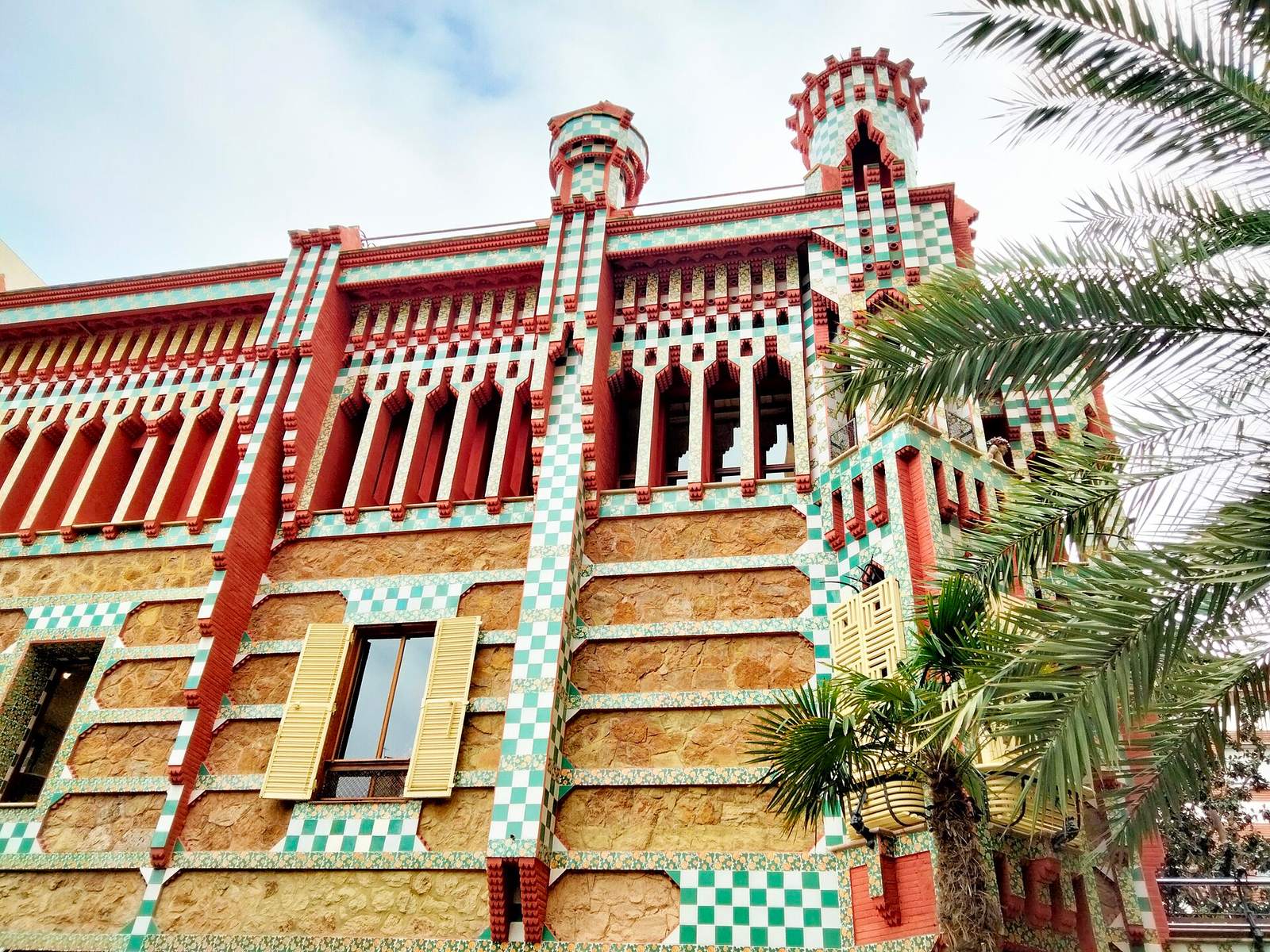 Casa Vicens's façade: red brick and ochre stone banded with green-and-white chequered tiles, small turrets and a row of slender columns under the roofline, beside a palm