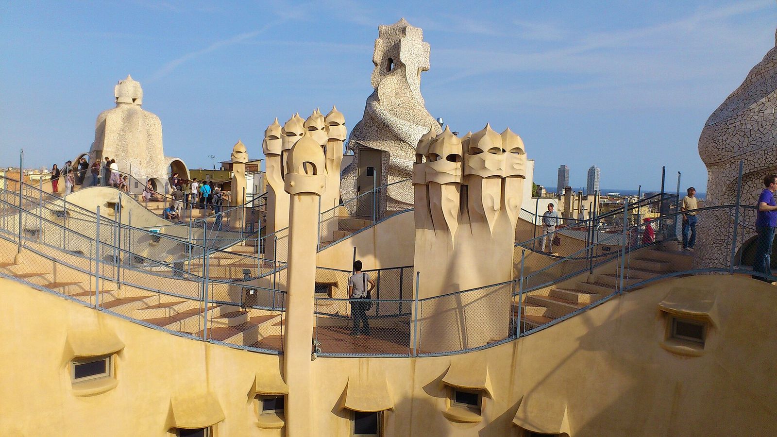 La Pedrera's rooftop in late-afternoon light: wavy stairways and sculpted chimney stacks, some covered in broken-tile mosaic, with visitors on the walkways