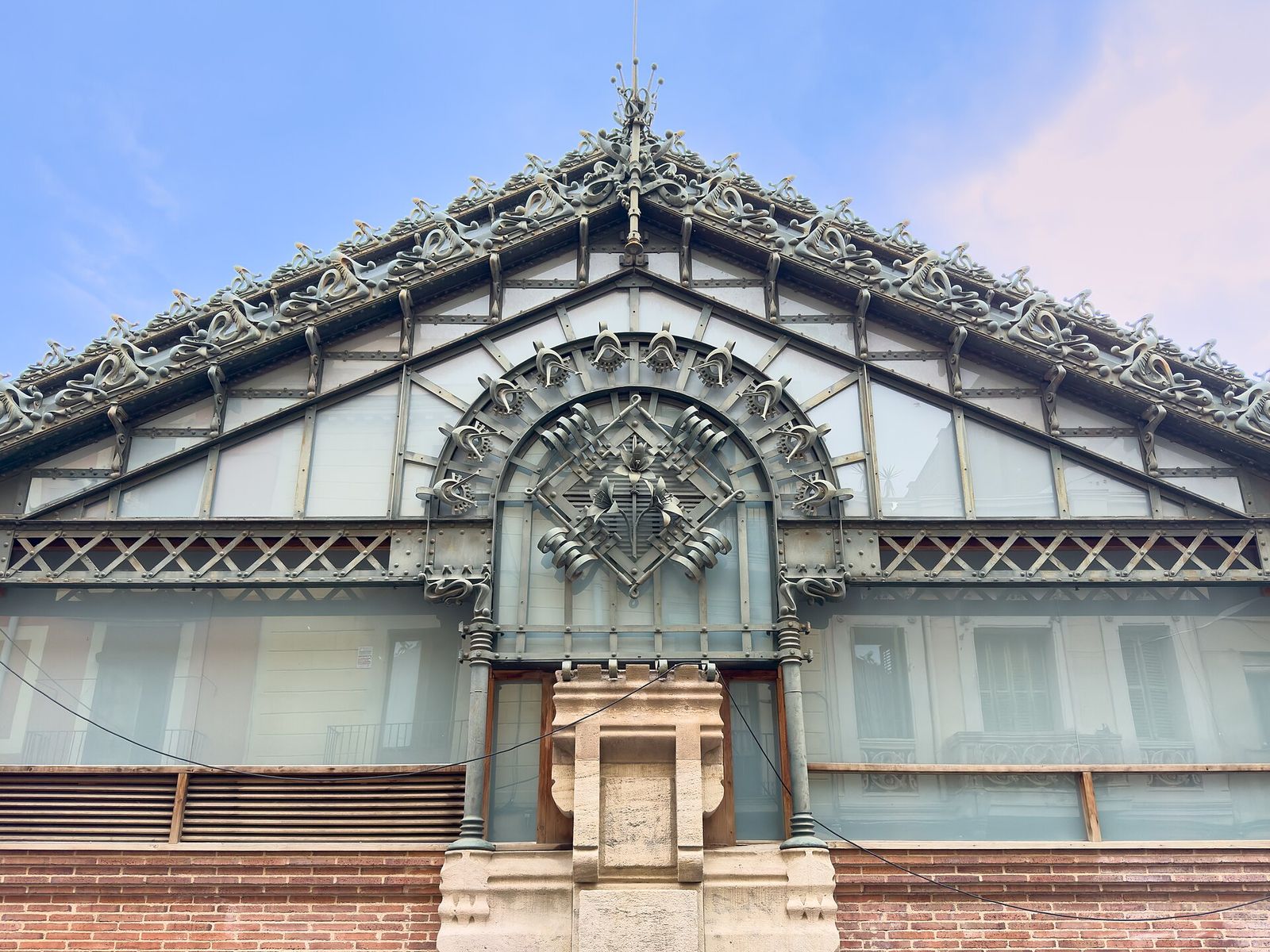 The iron-and-glass gable of the Mercat de la Llibertat, with ornate green ironwork and a coat of arms over a brick base, under a pale morning sky