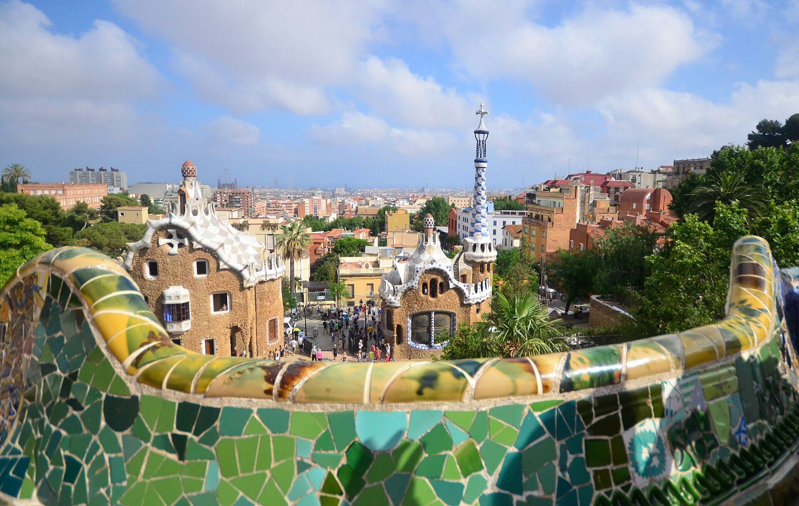 The curving serpentine bench on Park Güell's Nature Square, covered in green, yellow and blue broken-tile mosaic, looking down on the two gatehouse pavilions and the city