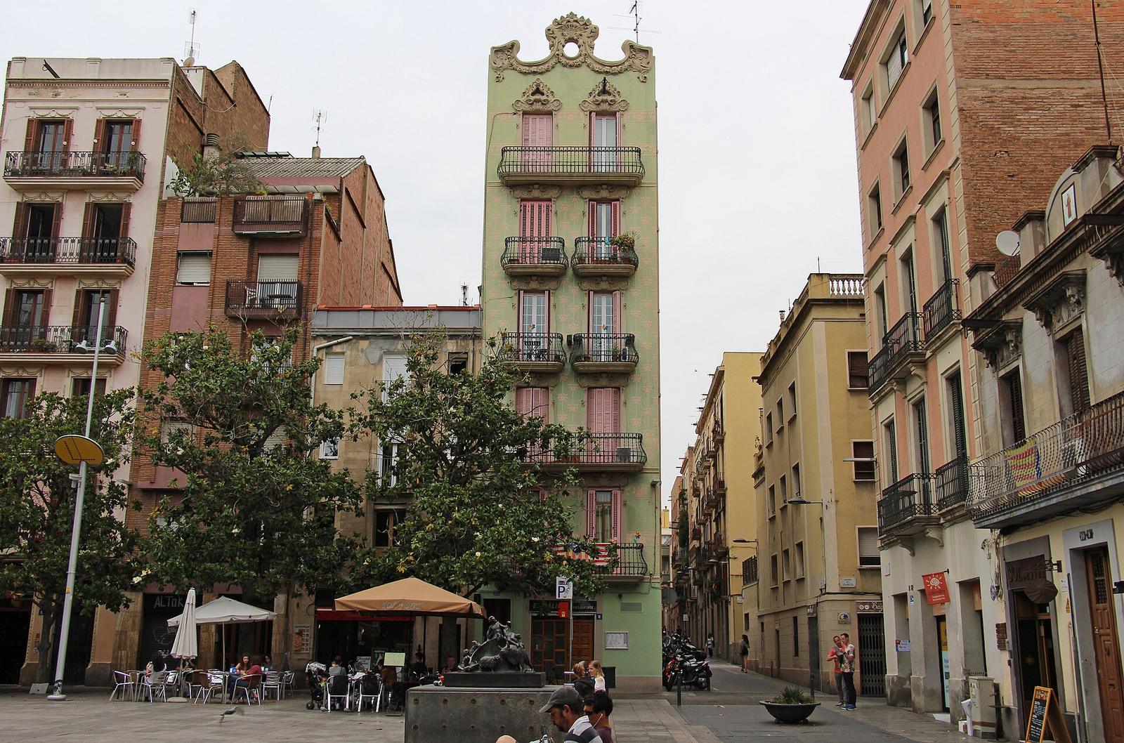 Plaça del Sol in Gràcia: café terraces with parasols beneath a narrow, pale-green Modernista building with curved balconies, among brick and painted façades