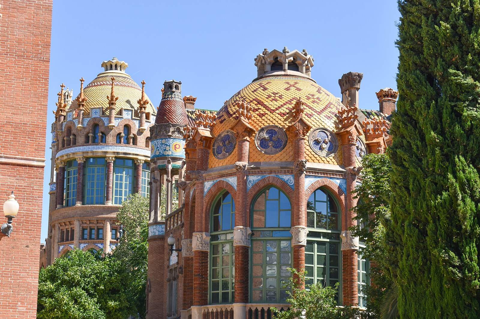 Two domed Modernista pavilions at Sant Pau, their cupolas covered in yellow and orange tiles above red-brick walls and tall green windows, beside tall cypress trees