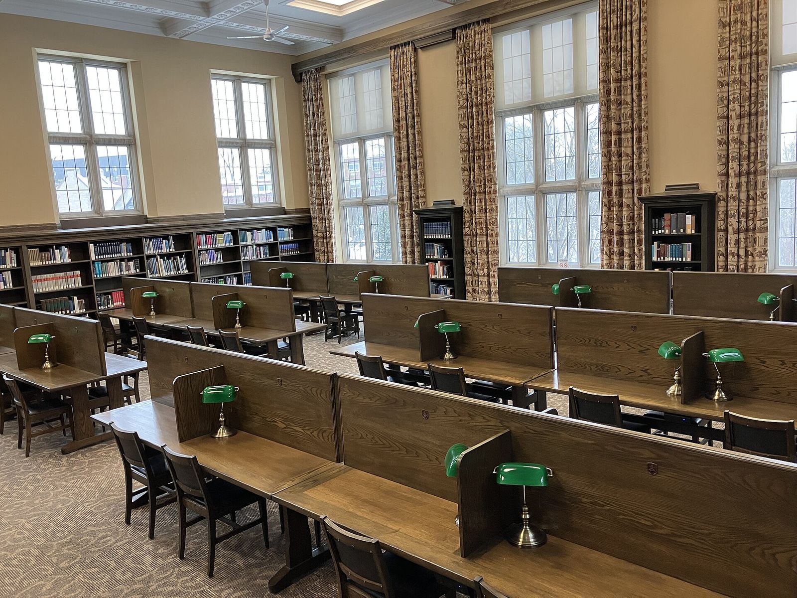 An empty quiet study room in a university library, with rows of wooden desks divided by low panels, a green banker's lamp at each place, bookshelves along the walls and tall windows