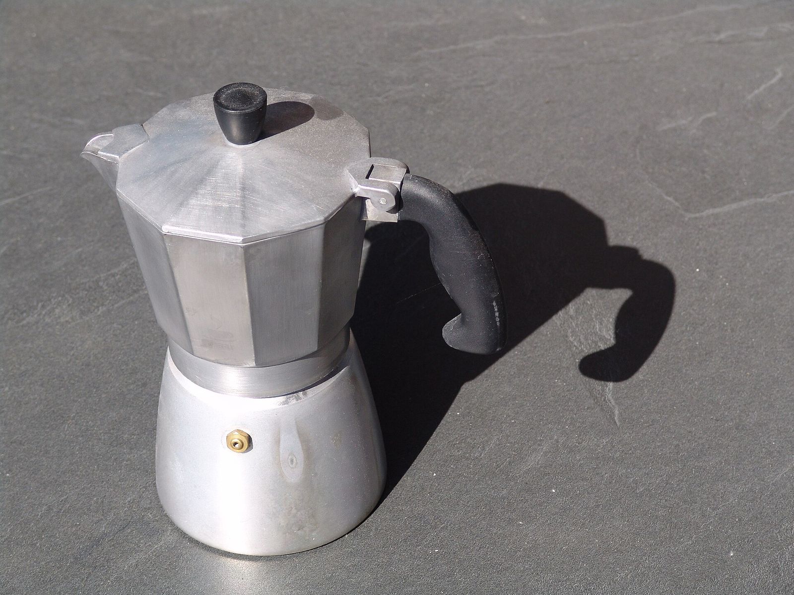 An aluminium stovetop moka pot with a faceted upper chamber, a black knob and a black handle, standing on grey slate in low sun that throws a long shadow of the handle