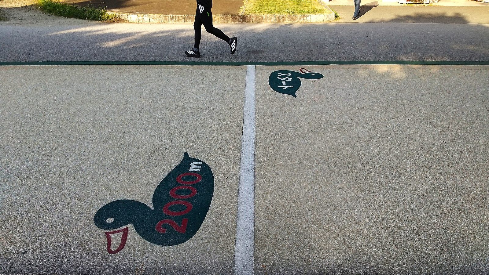 A paved park loop path painted with a green duck marked 2000 m and a second duck marked in Japanese, with the legs of a passing jogger at the top of the frame