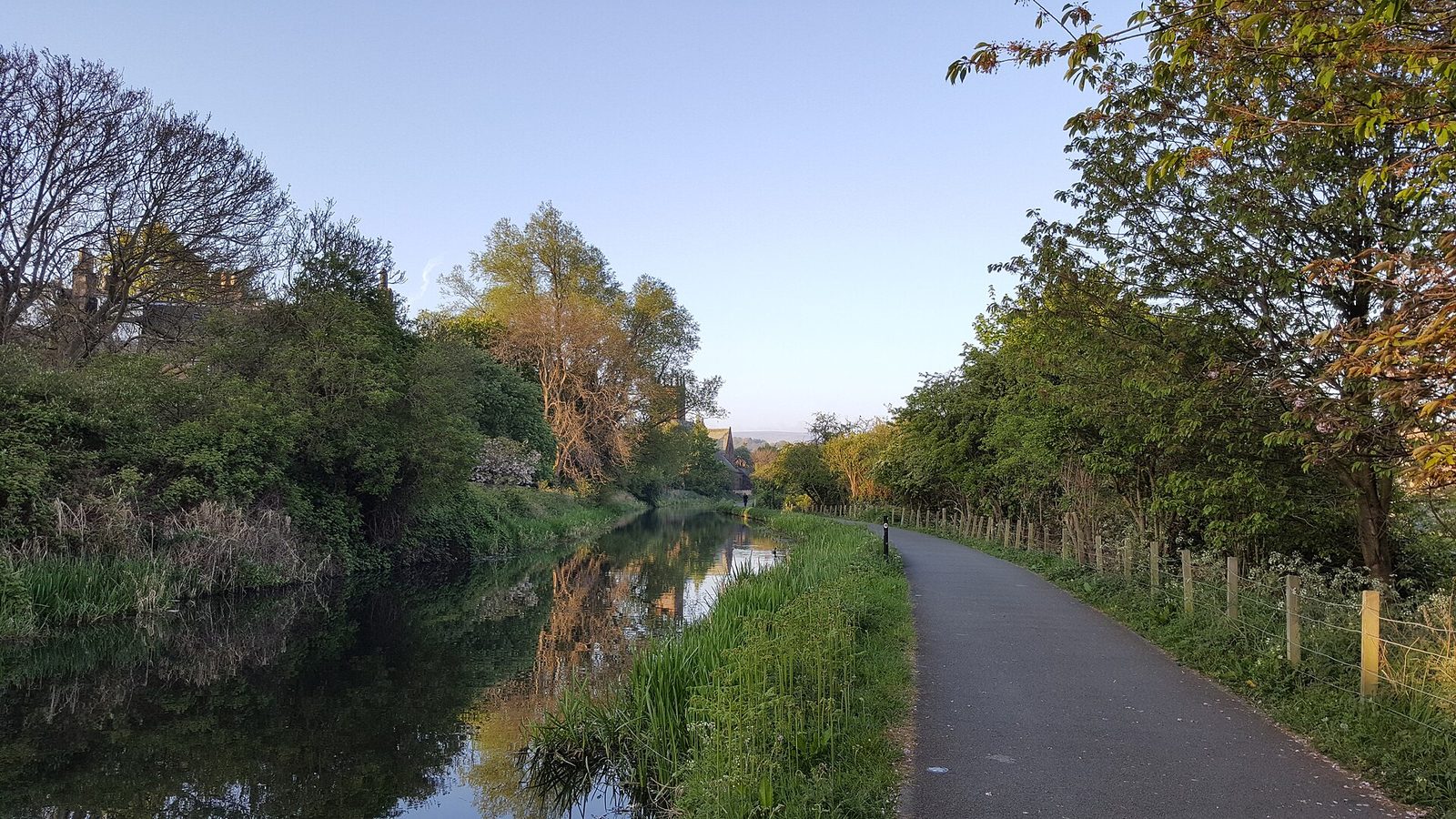 An empty paved towpath beside a still canal in early morning light, with trees reflected in the water and a wooden fence along the right-hand side
