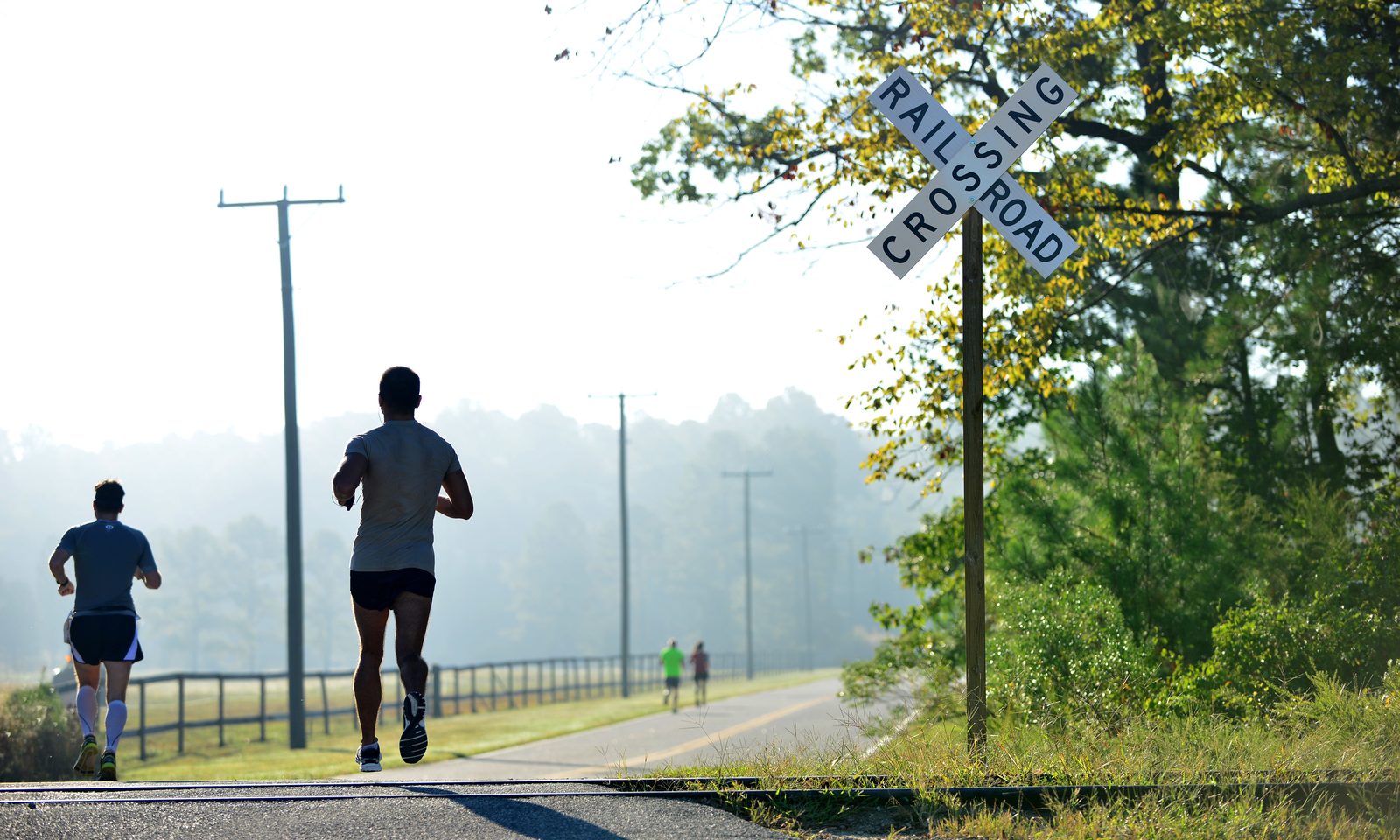 Two runners seen from behind in grey shirts and black shorts, running away along a paved path past a railroad crossing sign, with two more runners small in the morning haze ahead