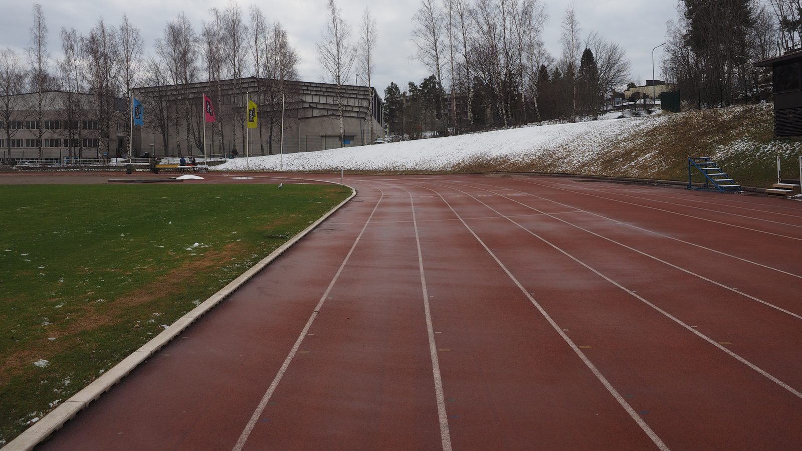 An empty red running track curving left past a strip of grass, with a snowy bank, bare birch trees and low buildings beyond under a grey sky