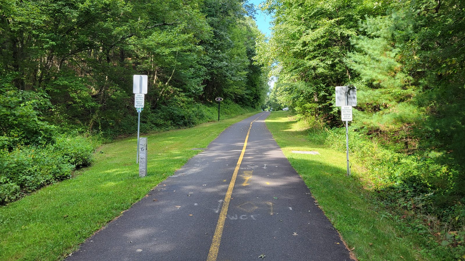 A paved rail trail with a yellow centre line running straight into green woods, with a granite mile 6 marker and trail signs on the grass verges