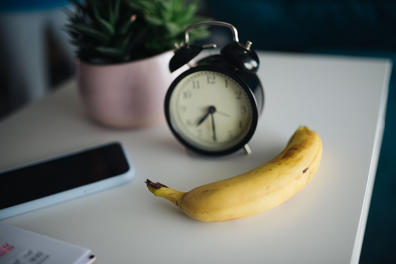 A black alarm clock on a white table next to a banana, with a smartphone lying face up with its screen off at the left edge and a potted succulent behind