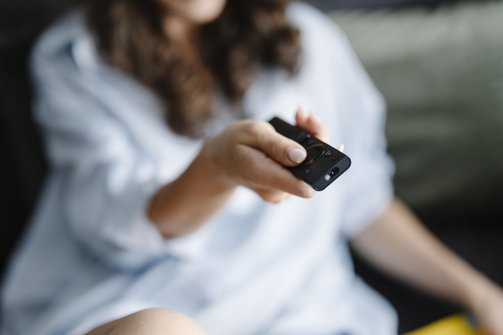 A person on a sofa, face out of frame, holding a black TV remote out towards the camera