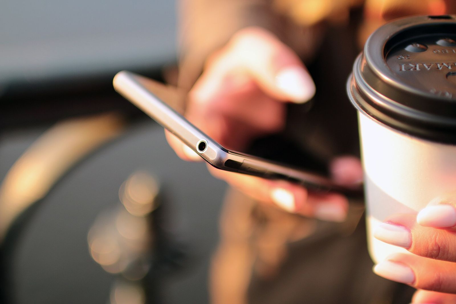 Close-up of one hand scrolling on a silver smartphone while the other holds a takeaway coffee cup, in warm light