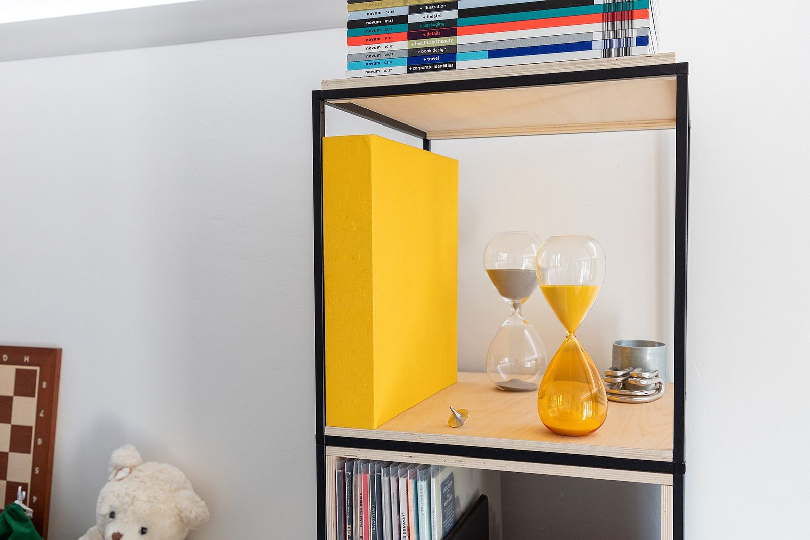 Two glass hourglasses on a wooden shelf, one with grey sand and one tinted yellow with yellow sand, beside a yellow box