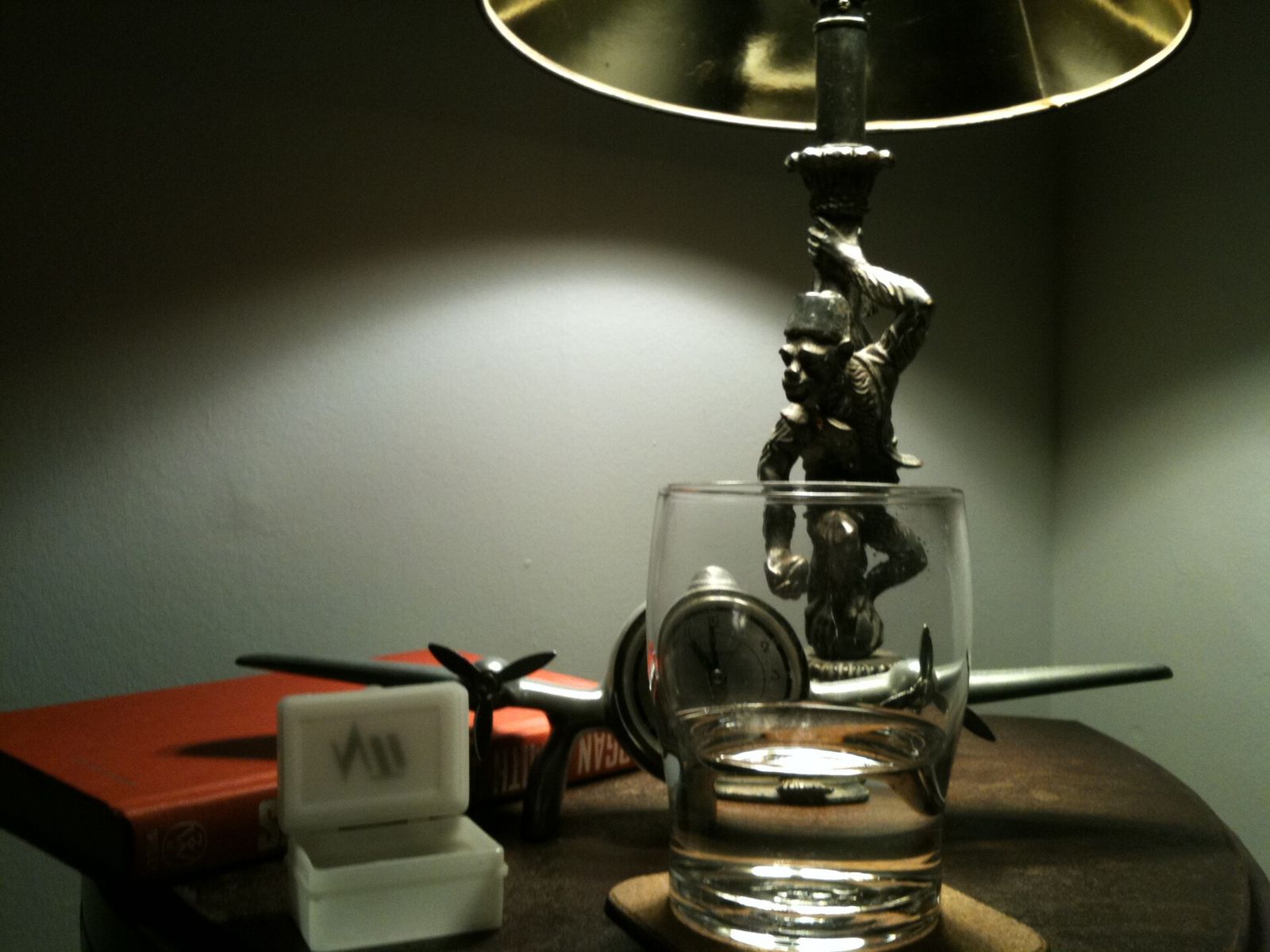 A dimly lit bedside table with a lamp, a glass of water, a small clock, a toy plane and a red book, and no phone
