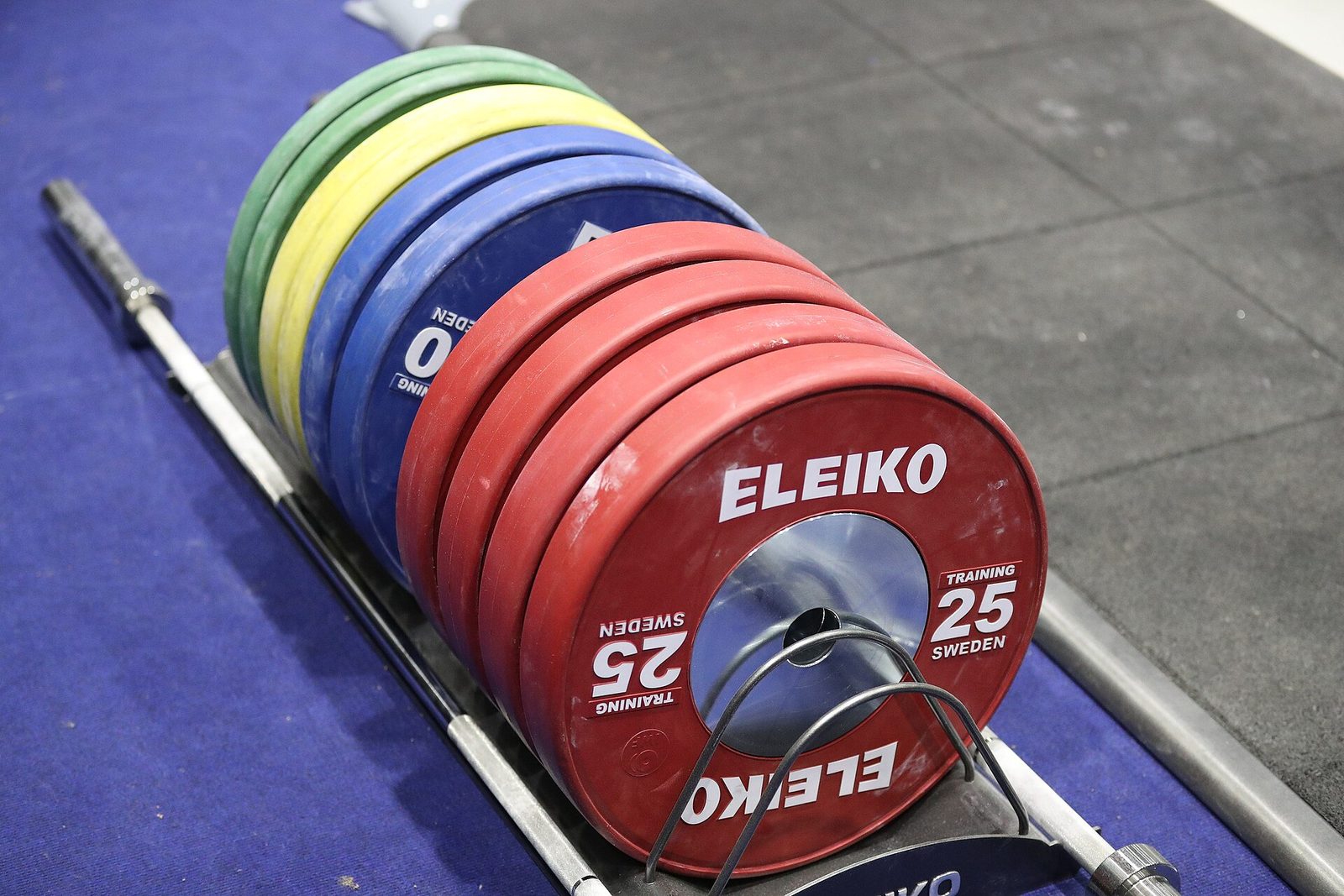 A rack of coloured bumper plates, red 25 kg plates at the front, beside a barbell lying on a blue lifting platform