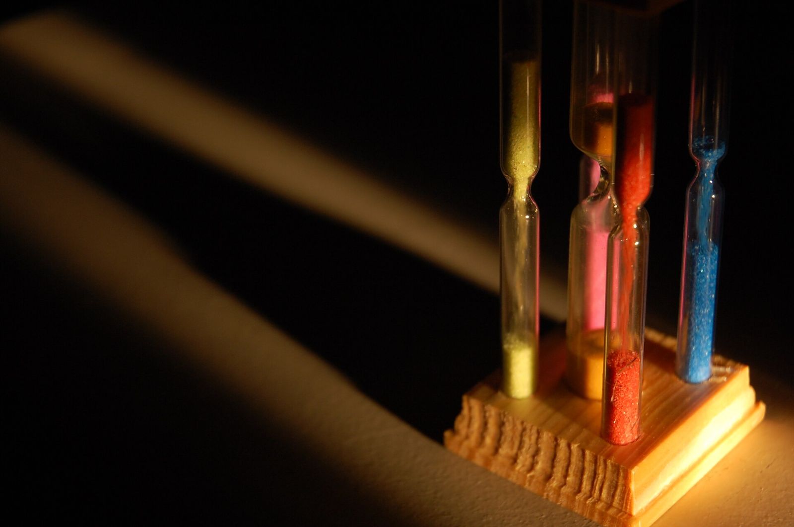 Four slim sand timers filled with yellow, pink, red and blue sand in a wooden stand, lit by a slanting beam of light in a dark room