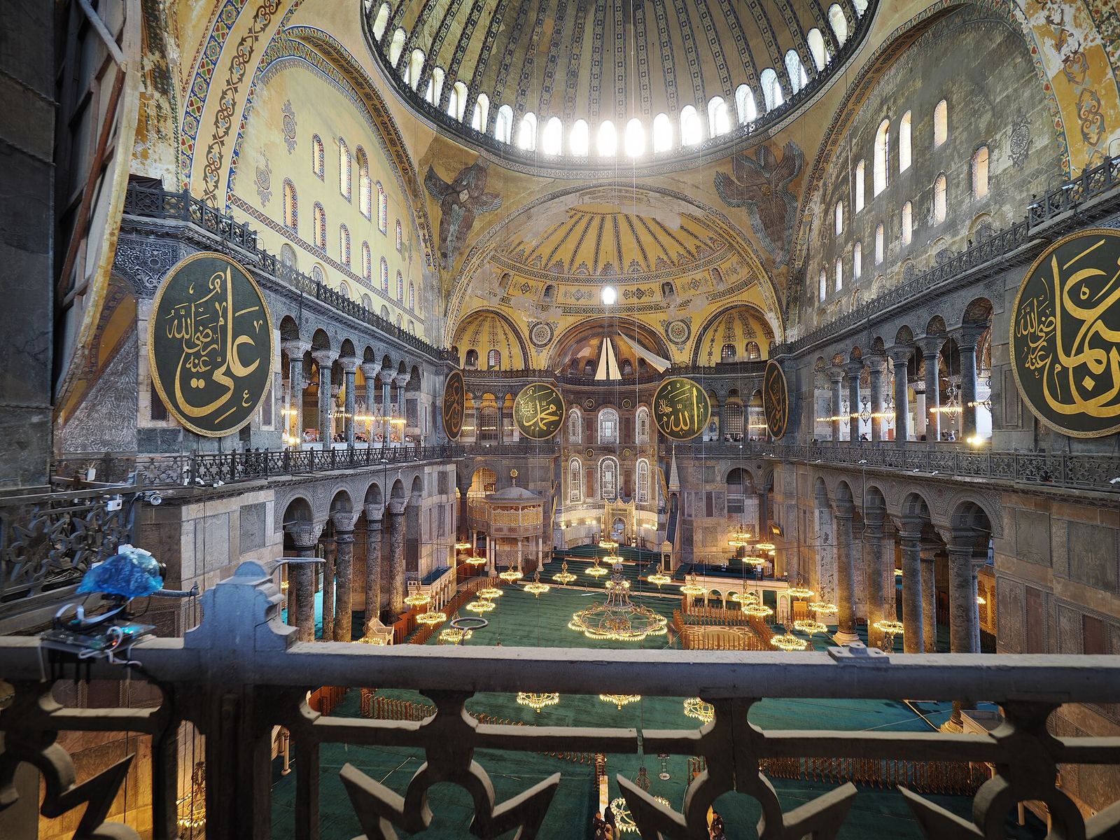 Hagia Sophia's nave seen from the upper gallery: the great dome, gold mosaics, round calligraphy panels and the carpeted prayer floor below