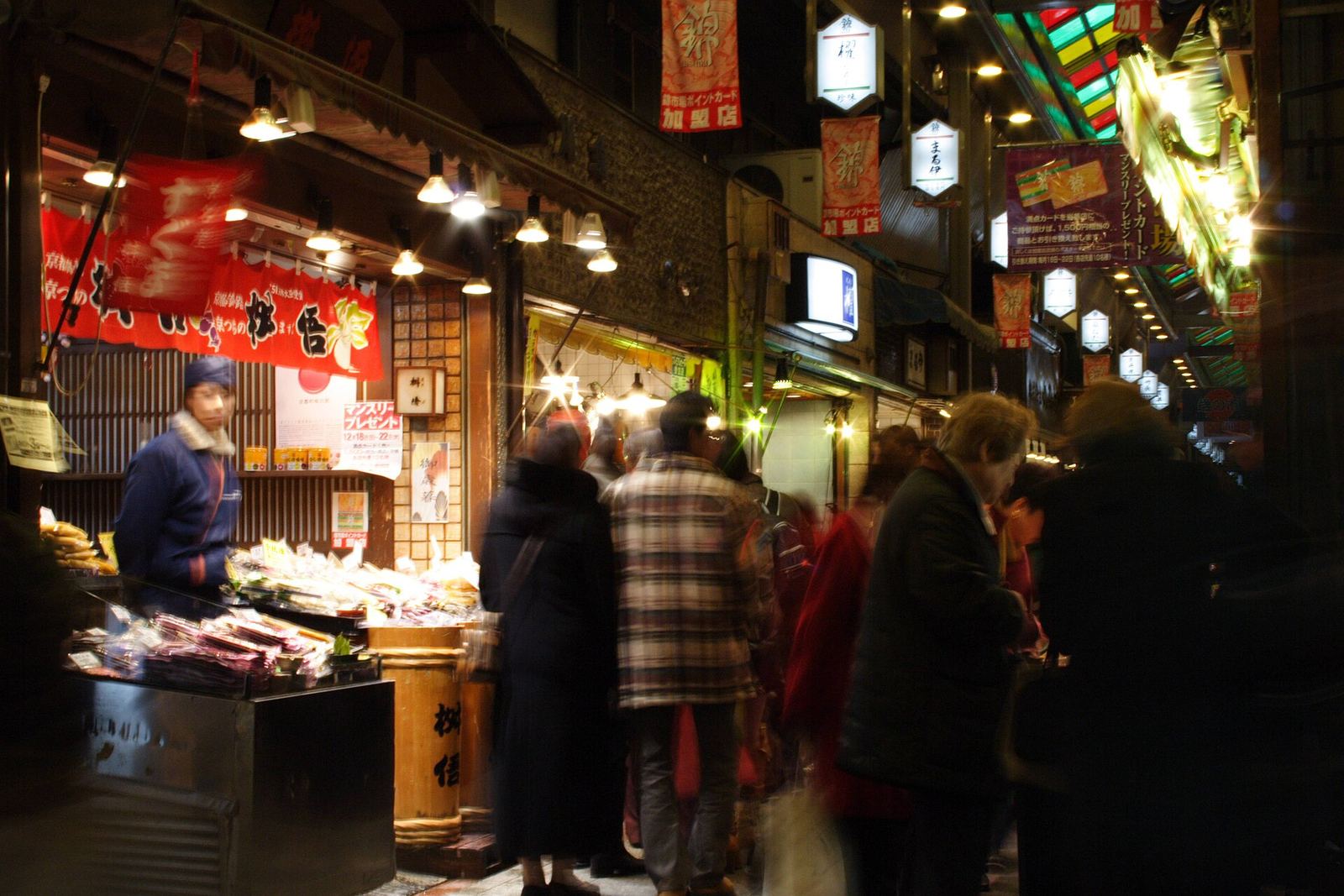 A brightly lit food stall in Nishiki Market piled with packaged goods, with shoppers passing under hanging signs