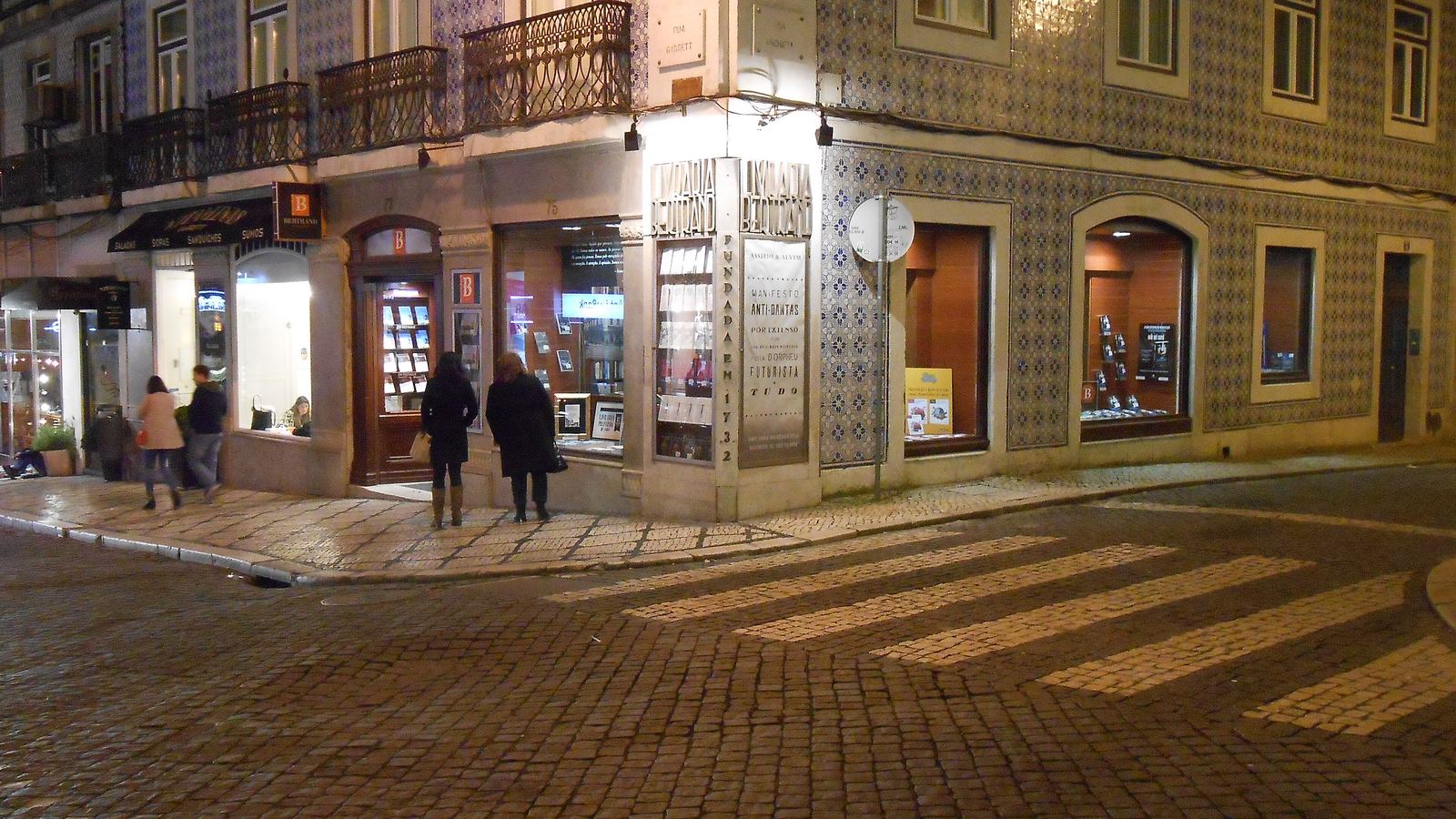 Livraria Bertrand's corner shopfront on Rua Garrett at night, its facade covered in blue-and-white tiles above lit windows, with a few people walking past