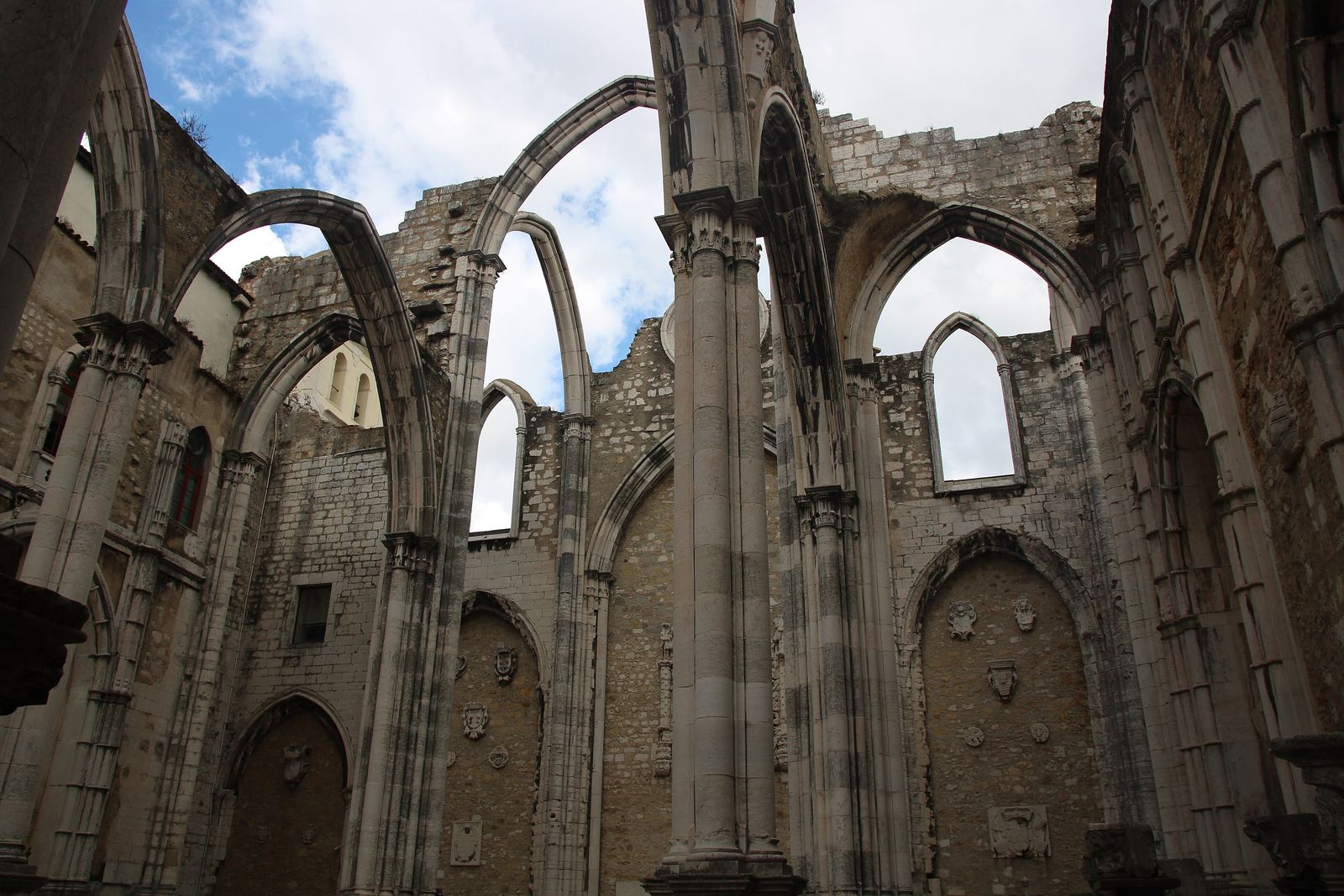 The roofless Gothic nave of the Convento do Carmo: tall pointed stone arches and columns standing open to a blue sky with white clouds