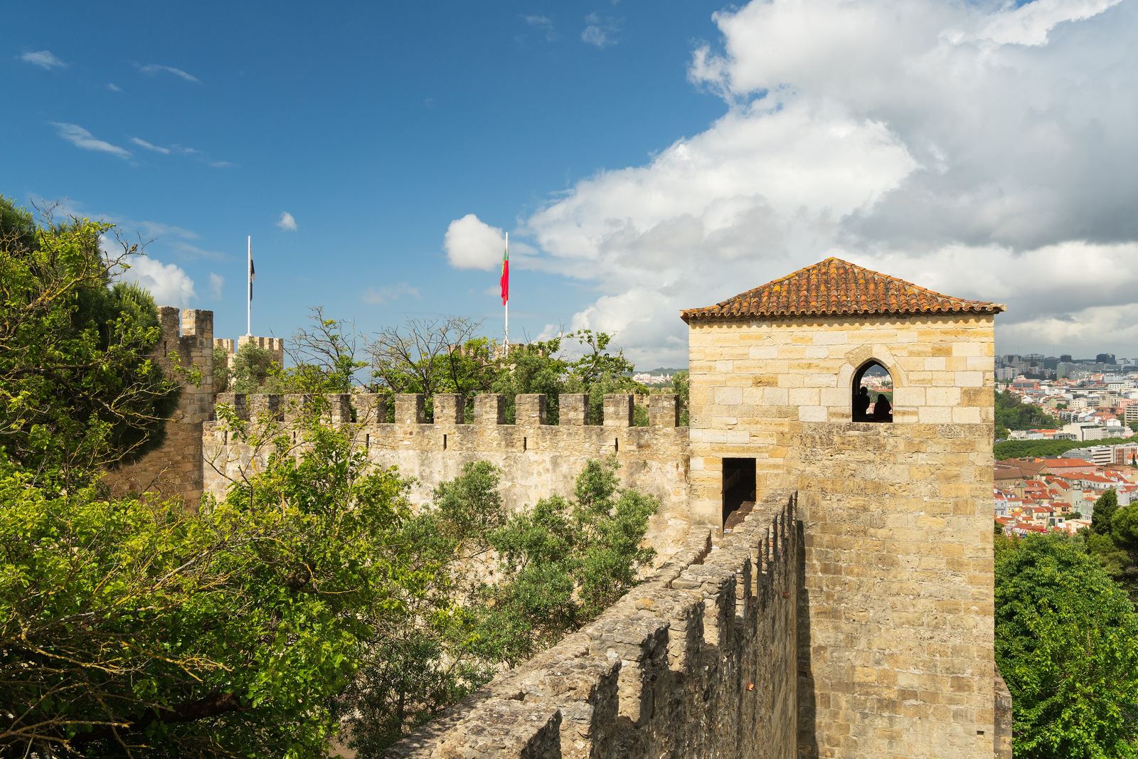 A crenellated stone wall and a square tower of the Castelo de São Jorge under a blue sky with white clouds, with trees and Lisbon's rooftops below