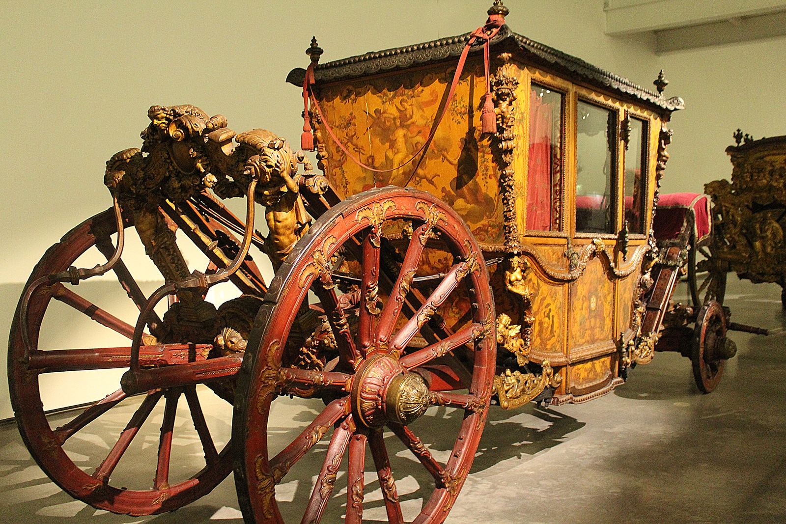 An ornate gilded coach with red-spoked wheels and carved figures on display inside the Museu Nacional dos Coches
