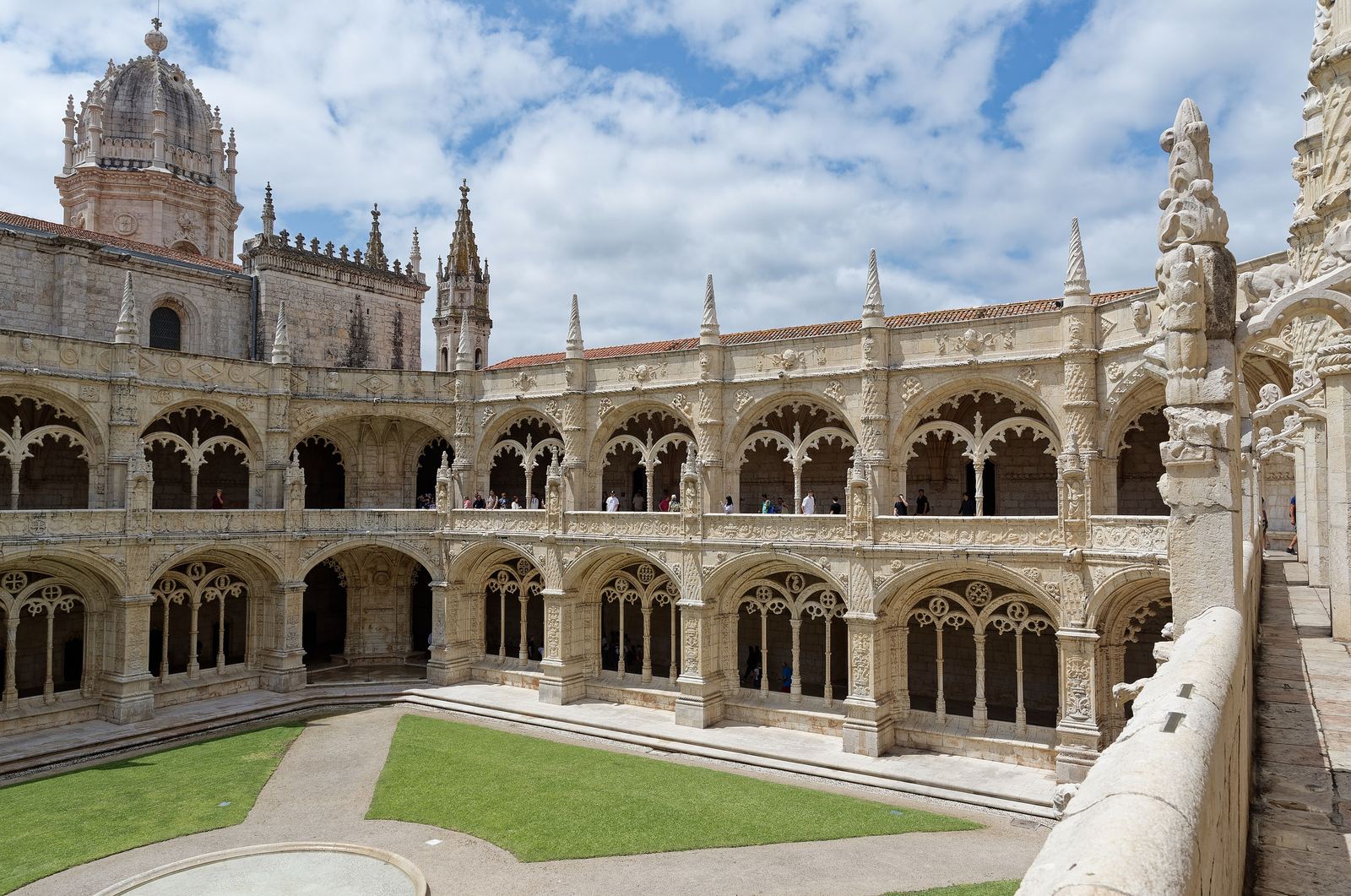 The two-storey cloister of the Jerónimos Monastery: carved limestone arches around a square lawn, with the church's domed bell tower rising behind under a partly cloudy sky