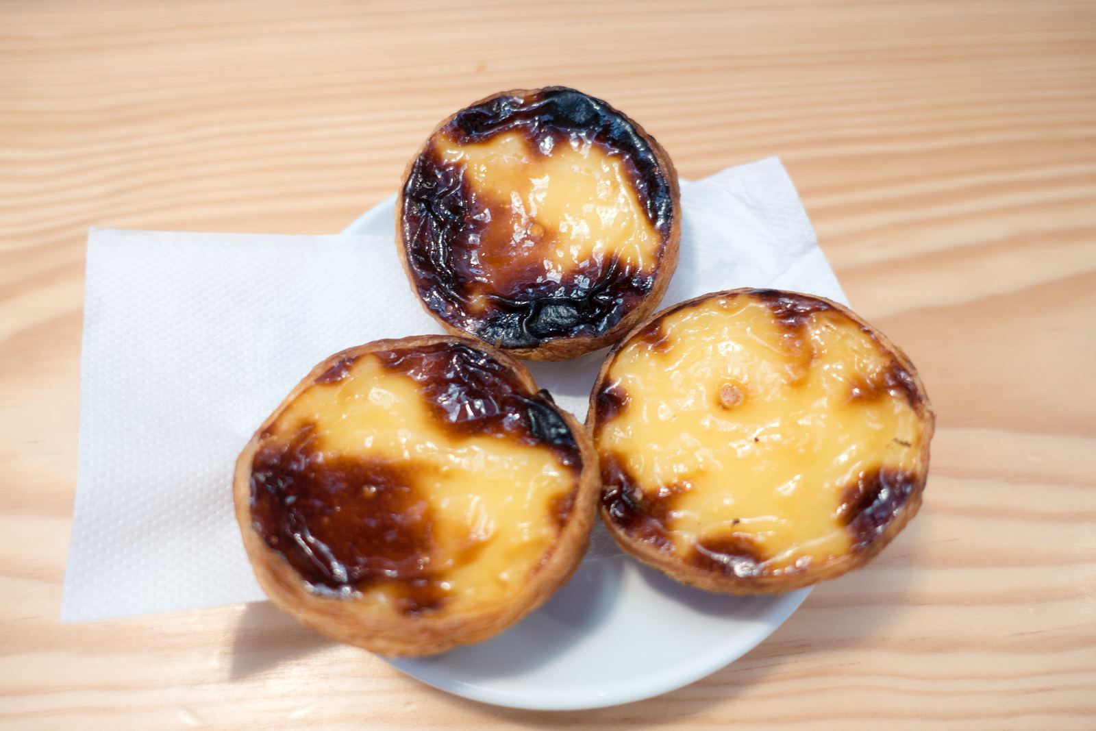 Three pastéis de nata with blistered, caramelised tops on a white plate and a paper napkin on a wooden counter
