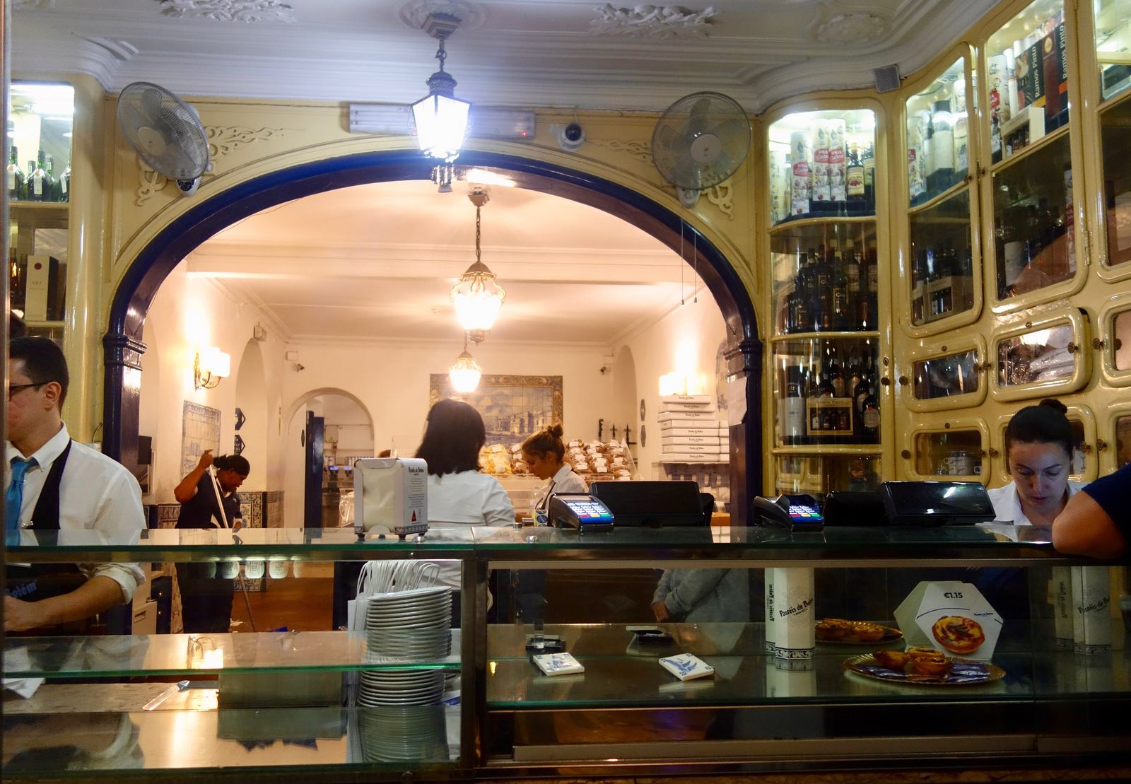 The takeaway counter at Pastéis de Belém: staff in white behind a glass counter, an arched doorway into a lit room behind them and shelves of boxes on either side