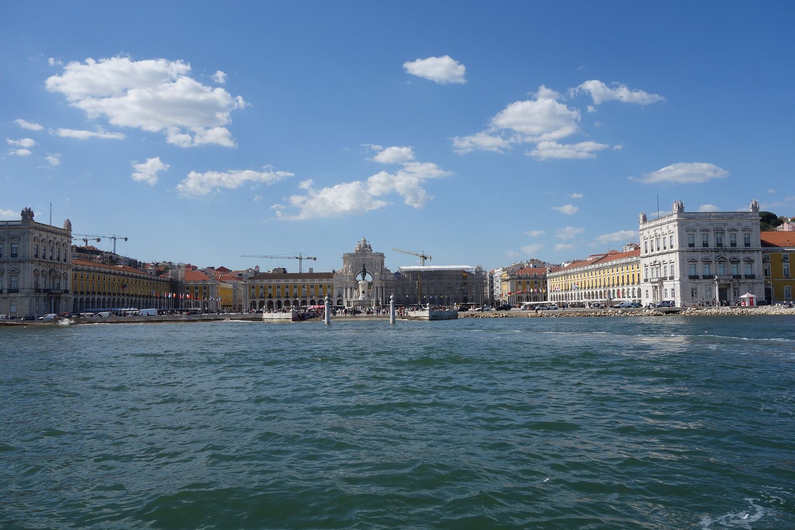Praça do Comércio seen from the Tagus: the long yellow arcaded buildings and the triumphal arch across the water under a blue sky with white clouds