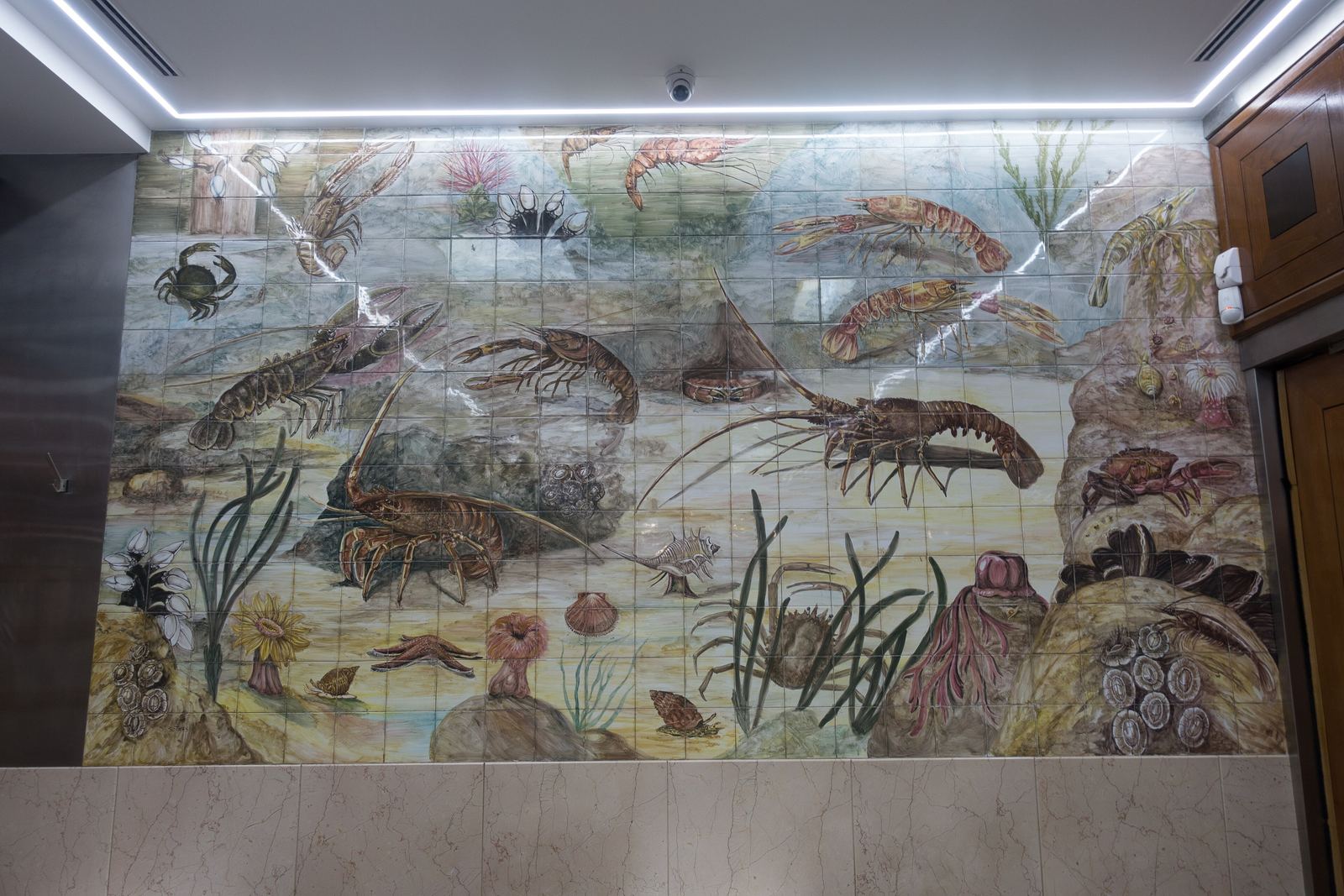 A large azulejo tile panel inside Cervejaria Ramiro painted with lobsters, prawns, crabs and fish among seaweed, lit by a strip of ceiling lights