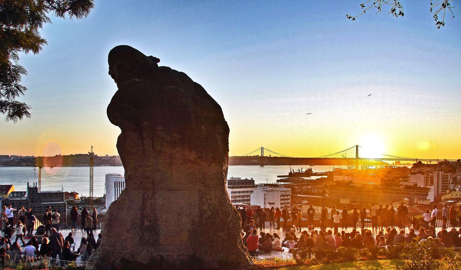 Sunset at the Miradouro de Santa Catarina: the silhouette of the Adamastor statue against an orange sky, the 25 de Abril bridge and the river behind, and a crowd sitting on the terrace