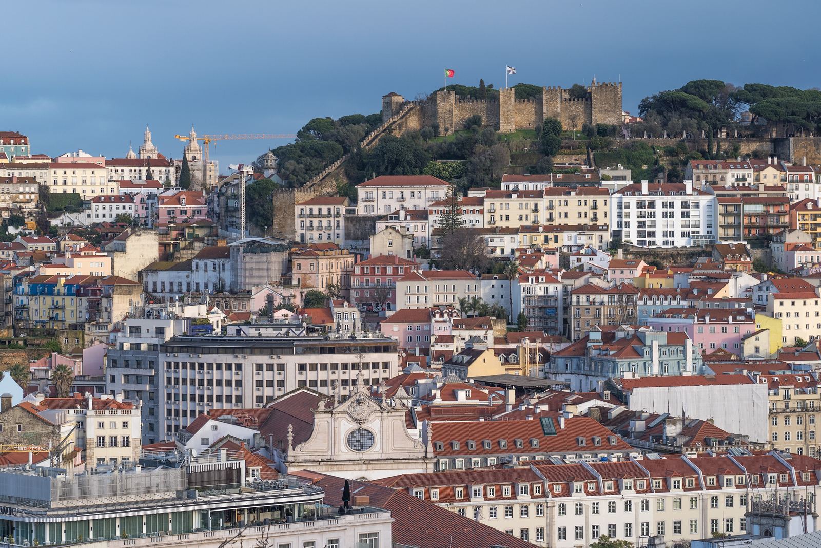 The view from the São Pedro de Alcântara viewpoint across the Baixa to the castle hill: tiers of pastel houses and red roofs rising to the walls of the Castelo de São Jorge