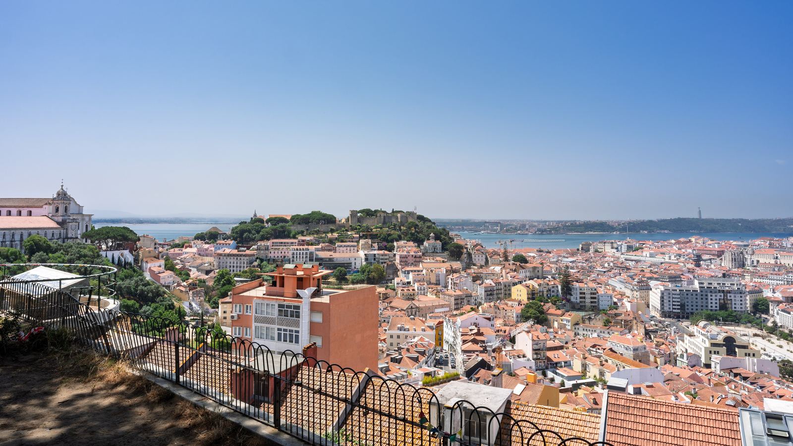A wide panorama of Lisbon from the Senhora do Monte viewpoint: the Castelo de São Jorge on its wooded hill at left, a sea of red roofs and pale buildings, and the Tagus across the horizon under a clear blue sky