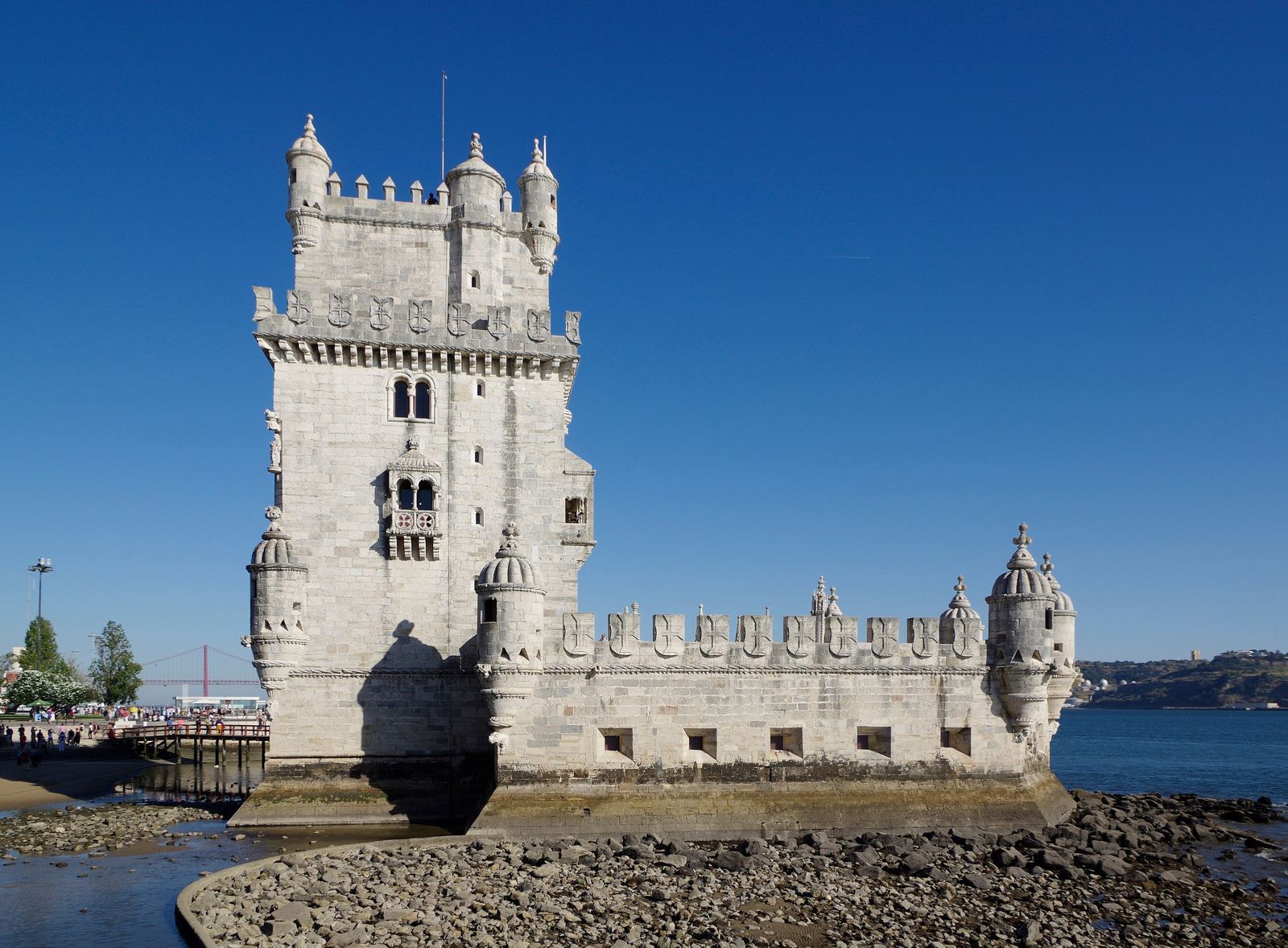 The limestone Torre de Belém with its turrets and battlements standing on rocks at the edge of the Tagus under a deep blue sky, with the red 25 de Abril bridge faint in the distance